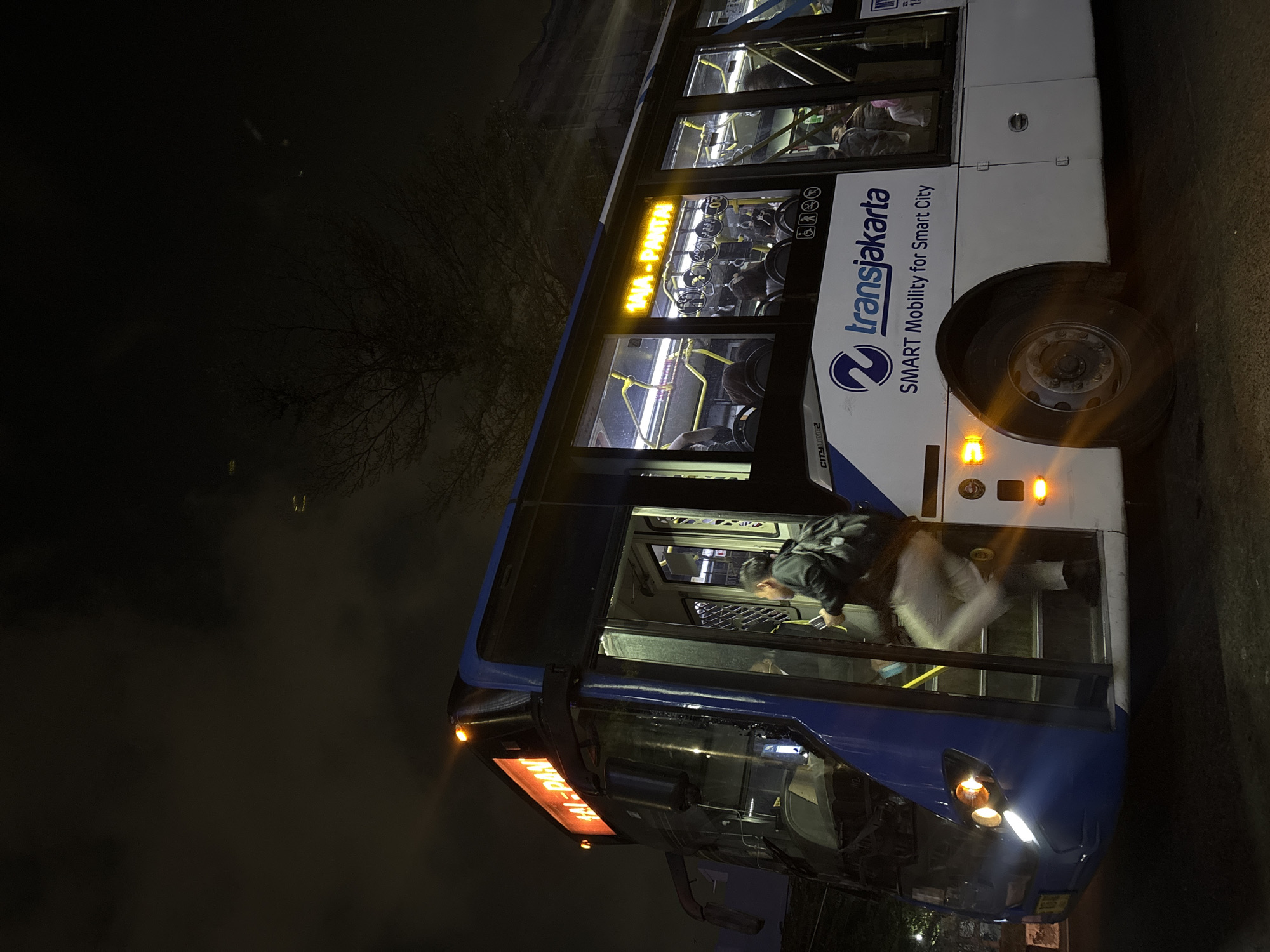 A low-angle, medium shot captures a Transjakarta bus at night, with the doors open and a person with a backpack ascending the stairs. The bus is blue and white, with the Transjakarta logo and the slogan "SMART Mobility for Smart City" visible on its side. The route destination, "TAN-PAN," is illuminated in orange LED lights at the front of the bus. The bus's headlights are on, casting a bright light on the street. The scene is set against a dark, cloudy sky, with a bare-branched tree visible in the upper right corner. The bus is likely parked on a street in North Jakarta, Indonesia, a common sight in the city's public transportation system. The interior of the bus is dimly lit, with yellow poles and seats visible through the windows. The person ascending the bus is wearing light-colored pants and a dark jacket, holding a phone. The overall impression is one of urban transit in the late hours.