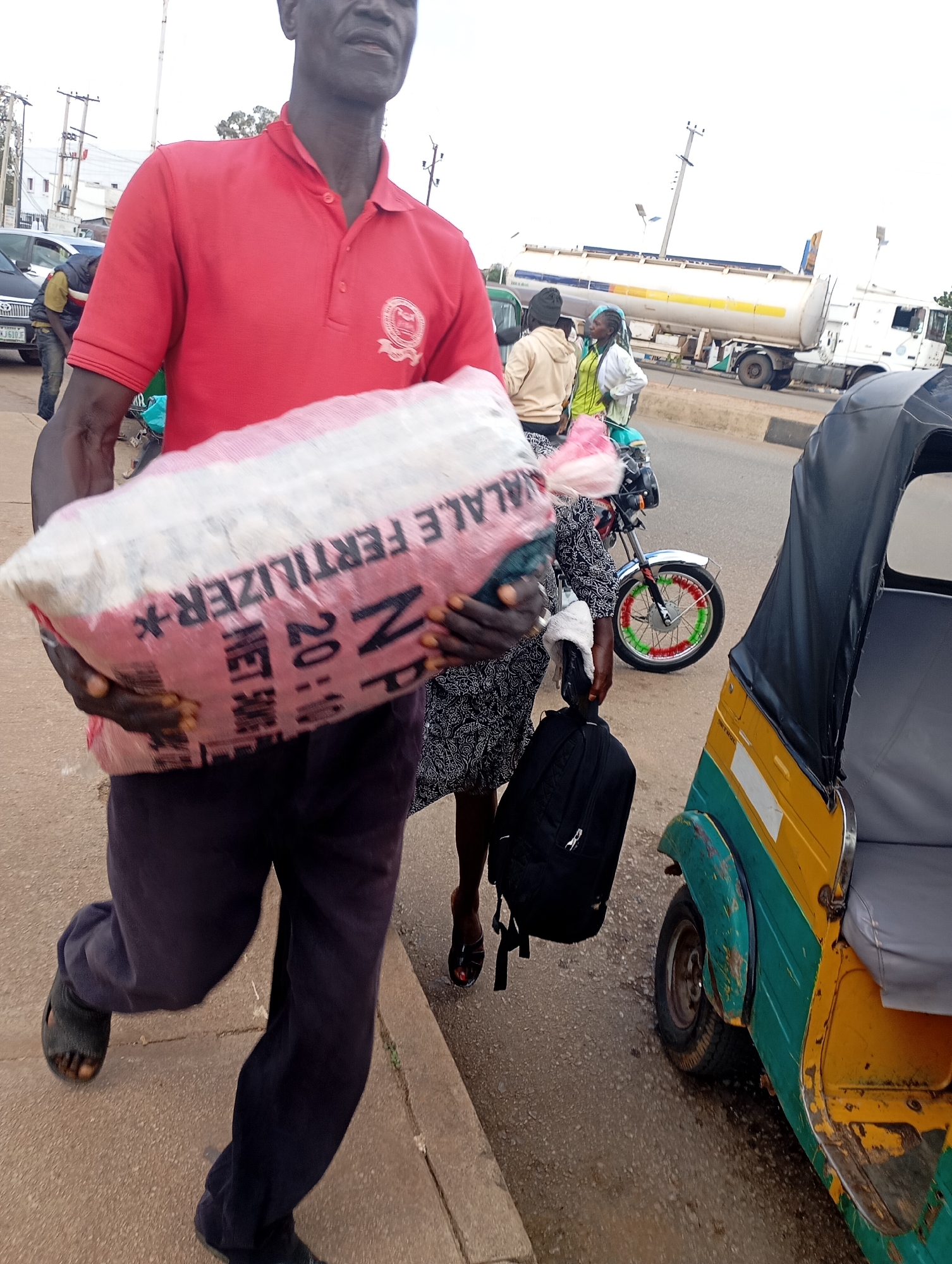 A man in a red polo shirt and dark pants is walking on a sidewalk in Gura Top, Nigeria. He is carrying a large bag of fertilizer. Behind him, a woman in a patterned dress and another person in a beige hoodie are walking. A tricycle, a motorcycle, a tanker truck, and cars are visible on the road. The scene appears to be a street in a Nigerian city.