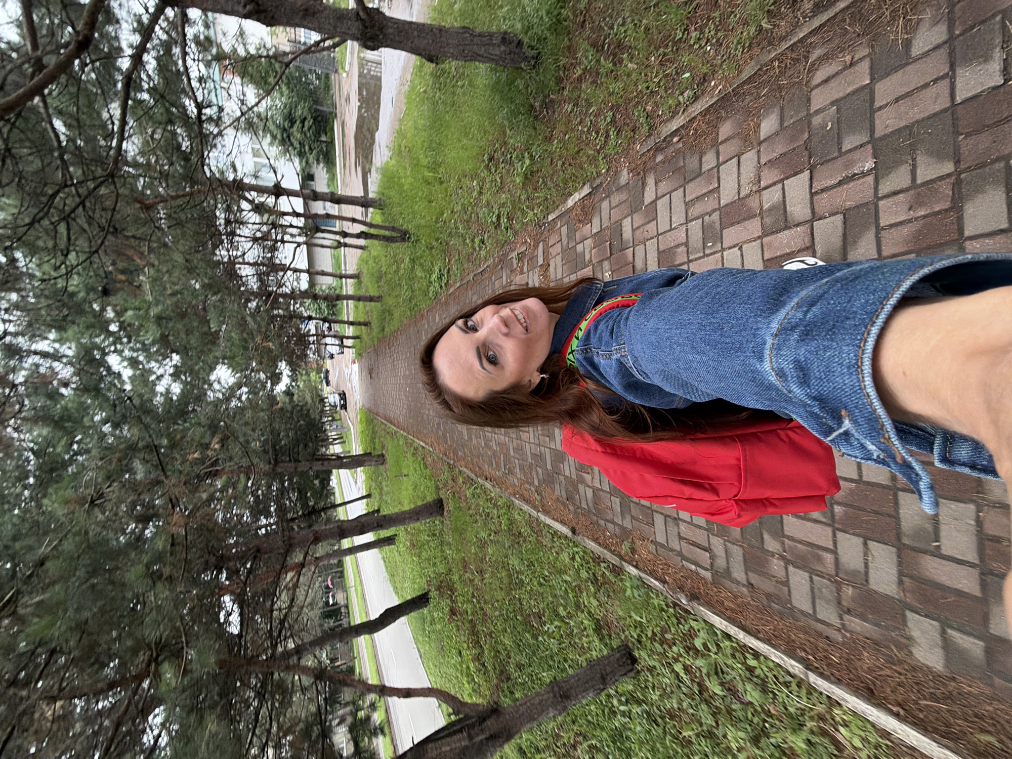 In Birobidzhan, Russia, a woman with long brown hair, wearing a denim jacket and a red backpack, is captured in a selfie. She is walking on a brick-paved path lined with trees on the left and grass on the right. A road with a few cars is visible on the left side of the path. The sky appears overcast. The scene is a typical urban street with greenery and trees.