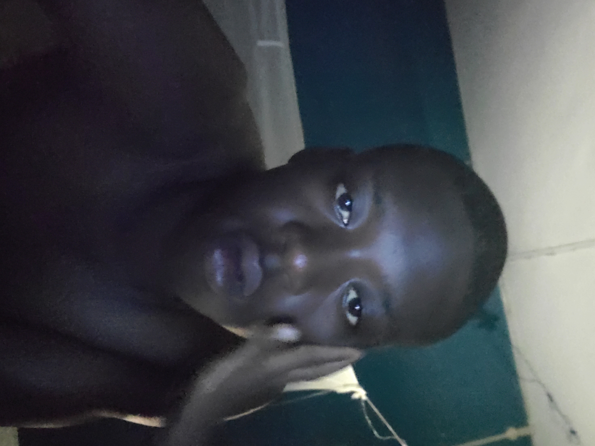 A person with dark skin is looking directly at the camera with their right hand touching the right side of their face. The background consists of two walls of differing colors. The location is Unknown, Unknown.