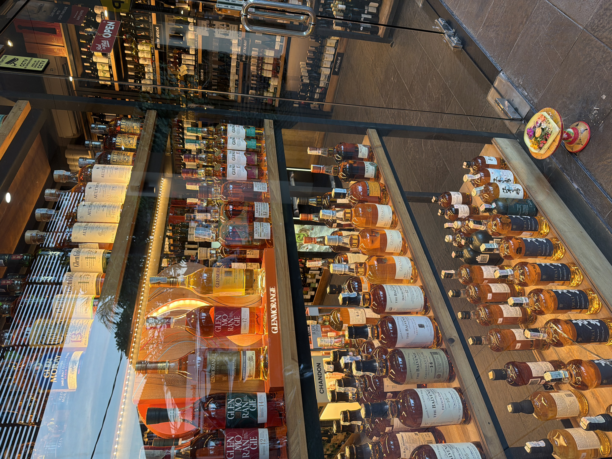 The image depicts a retail shop window displaying numerous bottles of various brands of single malt Scotch whisky. Prominent brands such as Glenmorangie, Glen Moray, Aultmore, and The Balvenie are visible. The bottles are arranged neatly on wooden shelves. Reflected in the glass are the interior shelves stocked with additional alcoholic beverages, along with exterior conditions including the presence of a bright blue sky. At the right corner of the window there are decorative floral arrangements on ornate stands. A sign in the reflection reads "OPEN", as well as several indicators related to CCTV surveillance, and country of origin signage. Based on the presence of "CCTV IN OPERATION" warnings, "EARN MILES WITH EVERY PURCHASE", and "AUSTRALIA, NEW ZEALAND, CANADA, SOUTH AFRICA" it's probable that this retail shop is located in a high-traffic tourist location such as the city of Kuta, Indonesia.