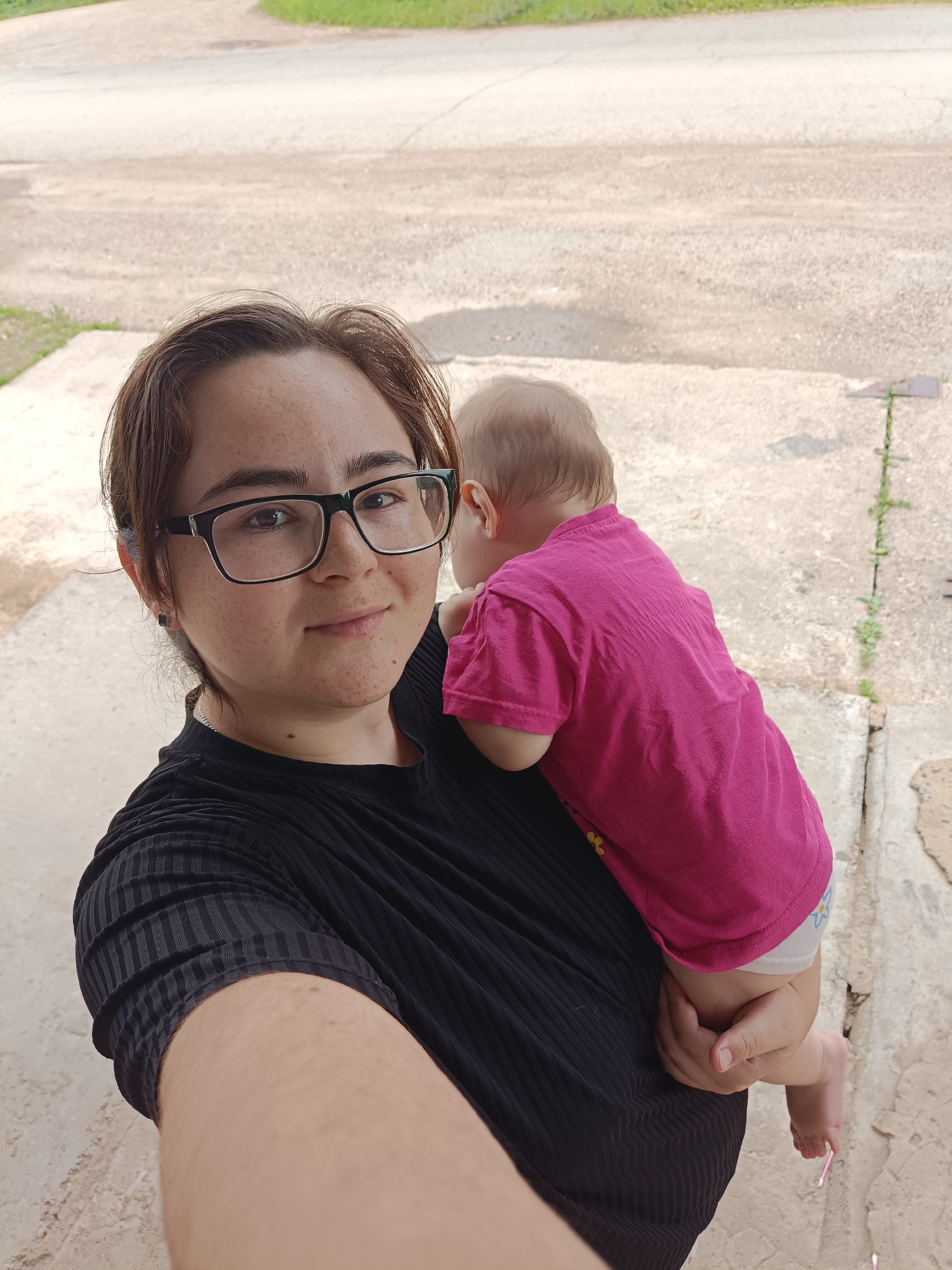 In a medium-shot, a young woman with short brown hair and glasses is taking a selfie while holding a baby. She is wearing a black top and the baby is wearing a pink t-shirt. The woman is smiling and looking at the camera. The baby is facing away from the camera, resting their head on the woman's shoulder. The background appears to be an outdoor, possibly urban setting, with a paved surface and some greenery visible. The overall impression is one of a casual, candid moment between a mother and child. There is no indication of the specific location in the city of Посад, Russia.
