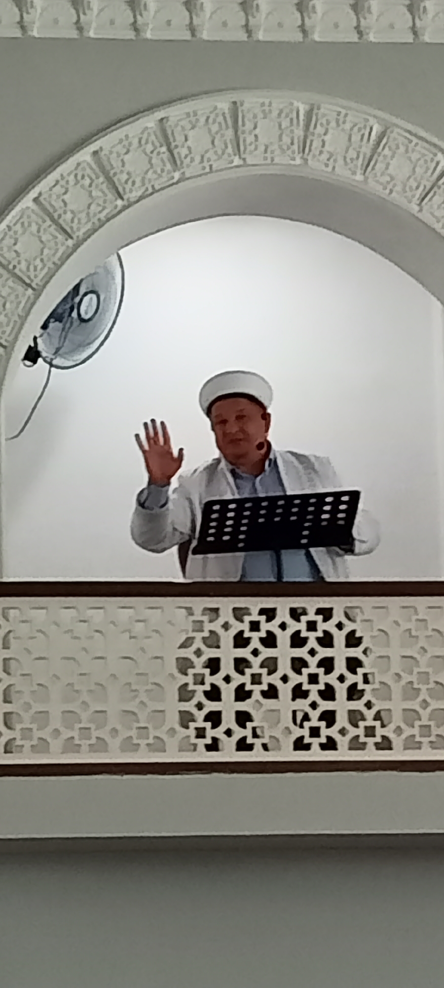 A man wearing a white Kufi and a light-colored jacket over a blue shirt stands behind a black music stand and waves with his right hand. He is positioned within a white arched alcove. A black fan is mounted on the white wall to the left of the man. A decorative white screen with a repeating geometric pattern runs horizontally in the foreground. The scene appears to be indoors, possibly in a mosque or a similar religious or community building in Almaty, Kazakhstan.