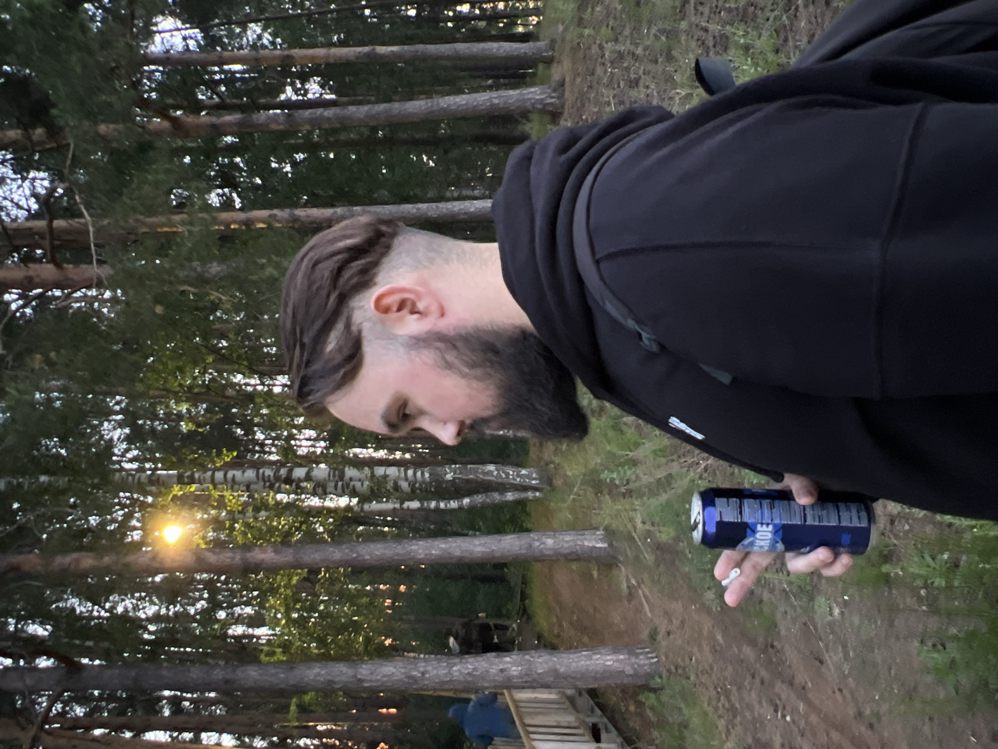 The image depicts a man in a forest environment, consistent with the location context of Chaikovsky, Russia. He is viewed in profile, facing left and looking downwards. The man has a full dark beard and a distinctive hairstyle with closely shaved sides and longer hair styled on top. He is wearing a dark, possibly black, hooded sweatshirt.
In his right hand, he holds a blue and silver aluminum beverage can. Partially visible text on the can, when viewed upright, appears to be "HOE" or similar, along with bar code details. Below the can, a white rolled cigarette is held between his fingers.
The scene is set in a dense wooded area, characterized by numerous tall trees, including slender pine trees and birch trees with their distinctive white bark. A bright, low-angled sunlight shines through the trees on the left side of the frame, indicating either early morning or late evening. The ground beneath the trees is a dirt path with scattered green foliage. In the distant background, a person wearing a blue hoodie is visible near a wooden structure, possibly made of pallets, and a ladder, suggesting a semi-developed or recreational area within the forest. The man appears to be in a moment of rest or contemplation in this natural setting.