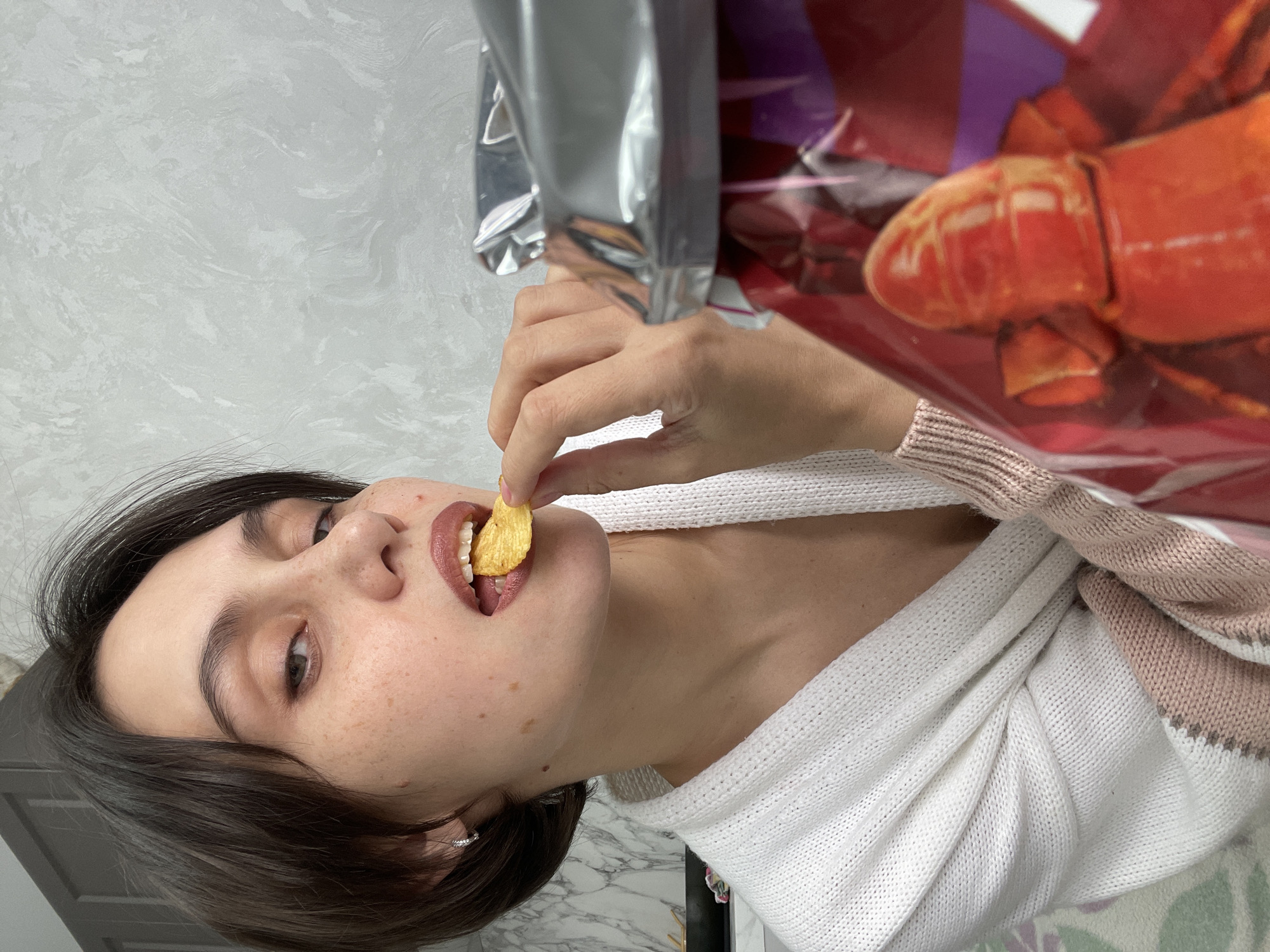 A young woman is holding a potato chip to her mouth. She is wearing a white sweater over a striped sweater, and a bag of chips is in the background. The background also includes a textured wall and a glimpse of what may be a marble surface. There is no information in the image indicating it was taken in Izhevsk, Russia.
