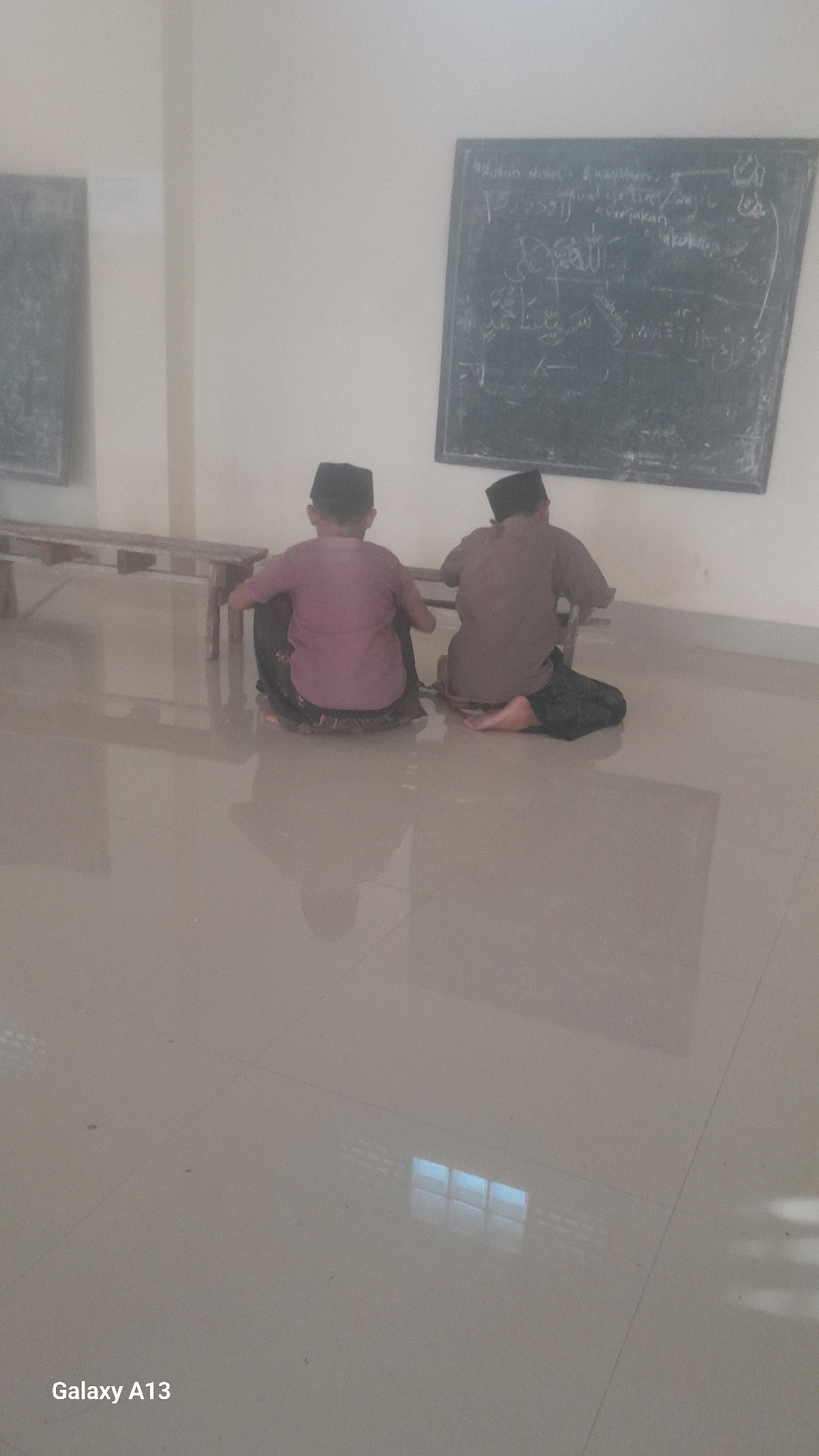 In Ngelo, Indonesia, two young boys are seen sitting on the floor in front of a chalkboard. The boy on the left wears a pink shirt and a patterned sarong, while the boy on the right wears a brown shirt and a patterned sarong. Both boys have black caps on their heads. The chalkboard behind them has writing on it, some of which appears to be in Arabic script. In the background, a wooden bench is visible. The floor is tiled and reflects light. The overall environment appears to be a classroom or a similar educational setting.