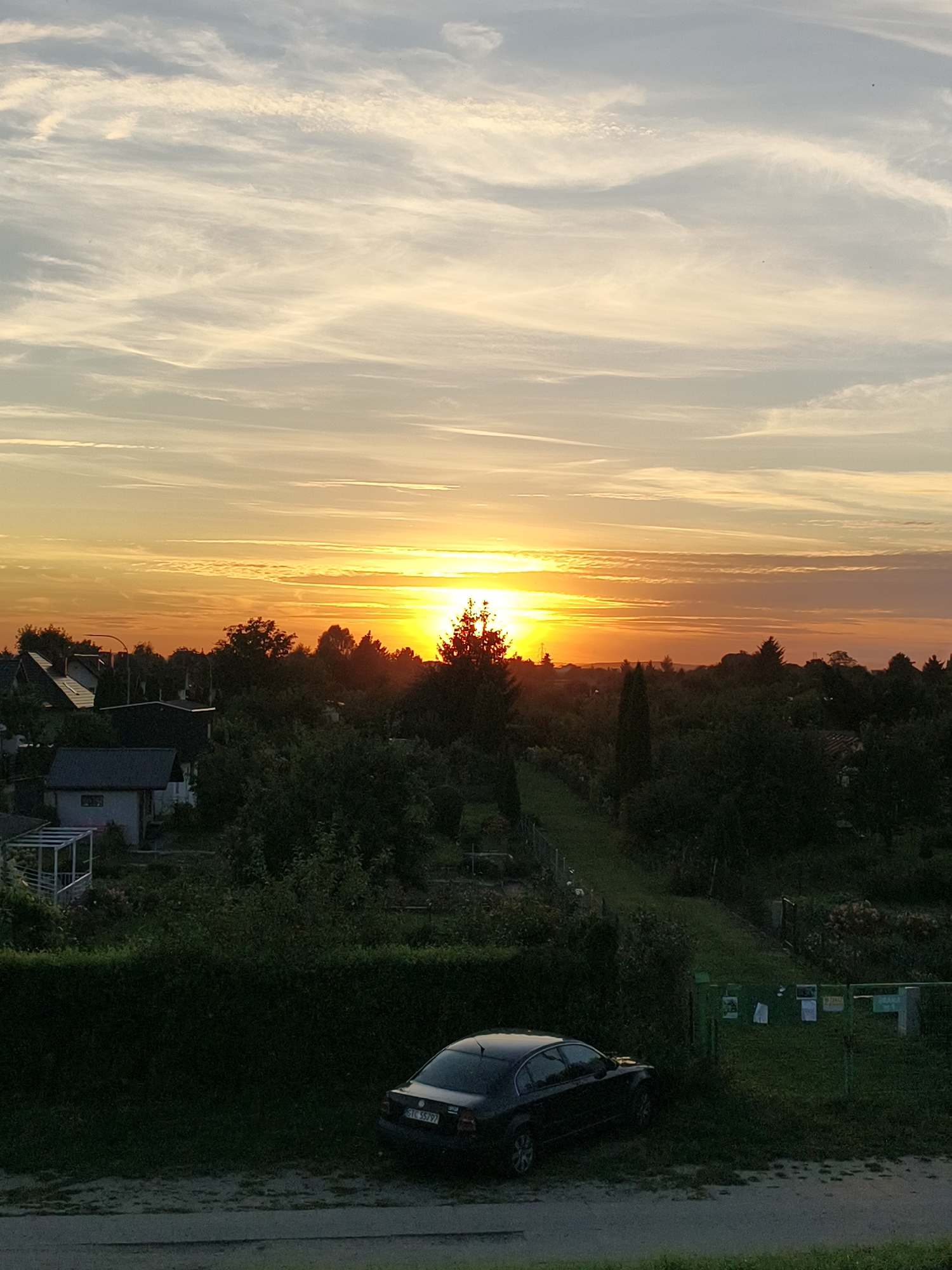 The image depicts a sunset over a suburban or rural area, likely on the outskirts of Tczew, Poland. The sky is filled with wispy clouds illuminated by the setting sun, which is a bright orange and yellow orb positioned towards the center-right horizon. Below the sky, there is a dense canopy of trees and vegetation, interspersed with several houses with dark roofs. A black sedan is parked on the left side of the frame, near a trimmed green hedge. A gravel path or driveway runs in front of the hedge, and beyond that, a road is visible. The scene suggests a quiet evening in a residential area.