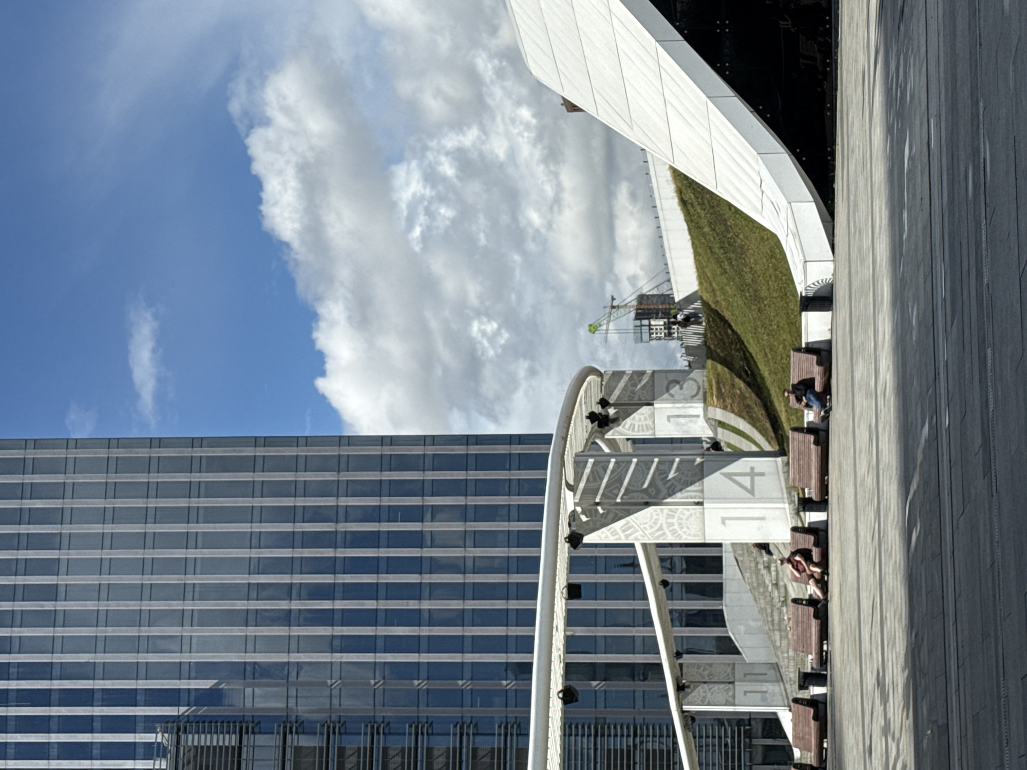 The image depicts a modern urban plaza in Moscow, Russia, likely part of a contemporary architectural complex. A towering glass and steel skyscraper dominates the left side of the frame, reflecting the bright blue sky and scattered white clouds. In the foreground, a paved plaza is lined with rows of dark wooden benches, some occupied by individuals. To the right, a futuristic, undulating white structure with a grassy slope behind it creates a striking contrast with the rigid lines of the skyscraper. A construction crane is visible in the background, suggesting ongoing development. Numbers "11" and "14" are visible on pillars supporting an overhead walkway, possibly indicating zones or sections within the plaza.