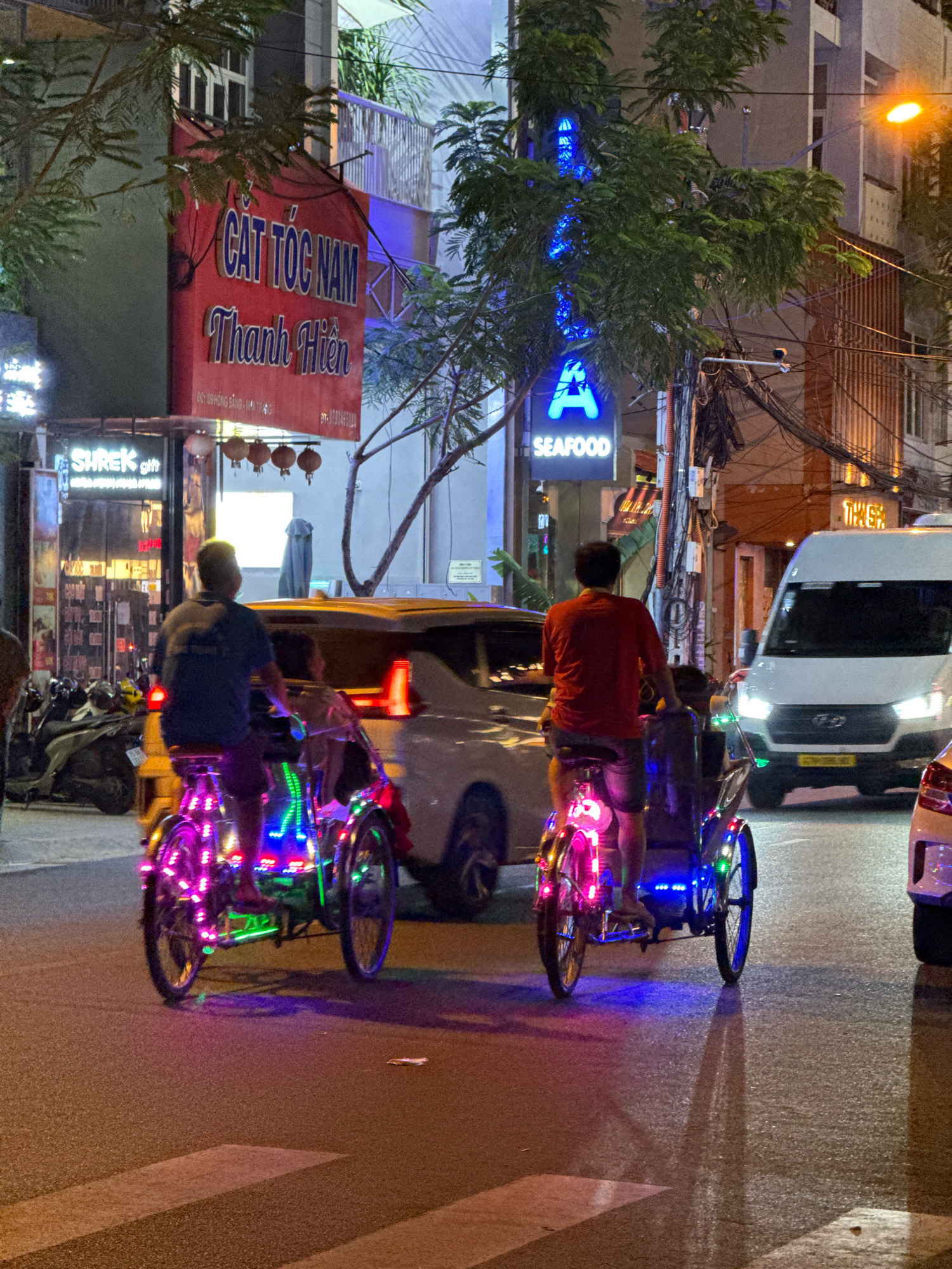 The image captures a nighttime street scene in Nha Trang, Vietnam. Two cyclos, decorated with colorful LED lights, are the central subjects. The cyclo on the left has a rider in a dark blue t-shirt and shorts, with a passenger behind them. The cyclo on the right has a rider in a red t-shirt and dark shorts, also with a passenger. Both cyclos are in motion on the street, illuminated by the vibrant lights.
In the background, various establishments are visible. A red banner with Vietnamese text, likely advertising a business, hangs prominently. Another sign displays "SEAFOOD" in blue neon letters. A white van is parked on the right side of the street, with its headlights on. The street is paved, and a pedestrian crossing is visible in the foreground. The overall atmosphere is lively, characteristic of a bustling urban environment at night.