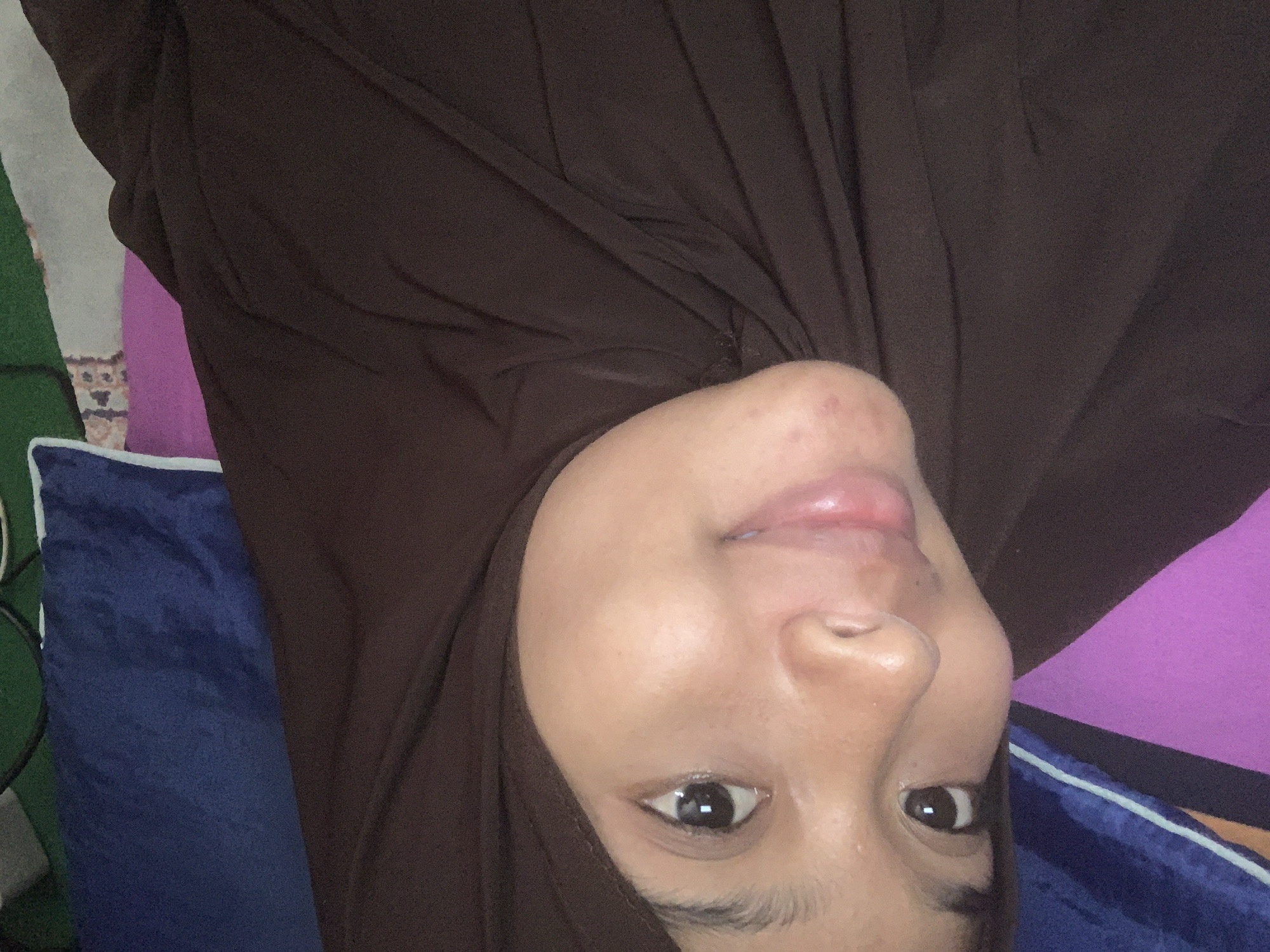 A close-up, head-and-shoulders selfie captures a young Indonesian woman in Sungai Meriam, Indonesia. She is wearing a brown hijab, which frames her face. Her skin appears clear, with a natural, dewy finish, and a few subtle blemishes are visible on her chin. Her eyes are dark and reflective, and she has a gentle, closed-mouth smile. The background is blurred but suggests a domestic setting, with draped fabric in shades of purple and blue, hinting at a bed or couch. The lighting is soft and diffused, illuminating her features and creating a warm, intimate atmosphere. The overall impression is one of natural beauty and serene self-possession.