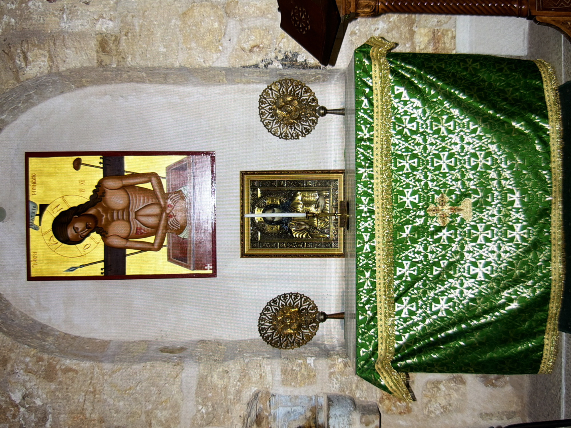 The image depicts an altar in what appears to be a religious setting, possibly a church or chapel, with stone walls and an arched ceiling. The altar is covered with a rich green cloth embroidered with a white, geometric cross pattern and trimmed with gold fringe. On the altar, from left to right, are two ornate, circular, golden objects with intricate designs, a framed icon of Jesus Christ with a halo and wearing a crown of thorns, and a lit candle in a golden holder. Above the altar, a large icon of Christ on the cross is displayed on the wall, depicting him with wounds and seated in a sarcophagus-like structure. To the right of the altar, a wooden lectern with a book is visible. The overall impression is one of a sacred space adorned for worship. The image's metadata does not specify a location in Kyiv, Ukraine, but the context suggests a religious building.