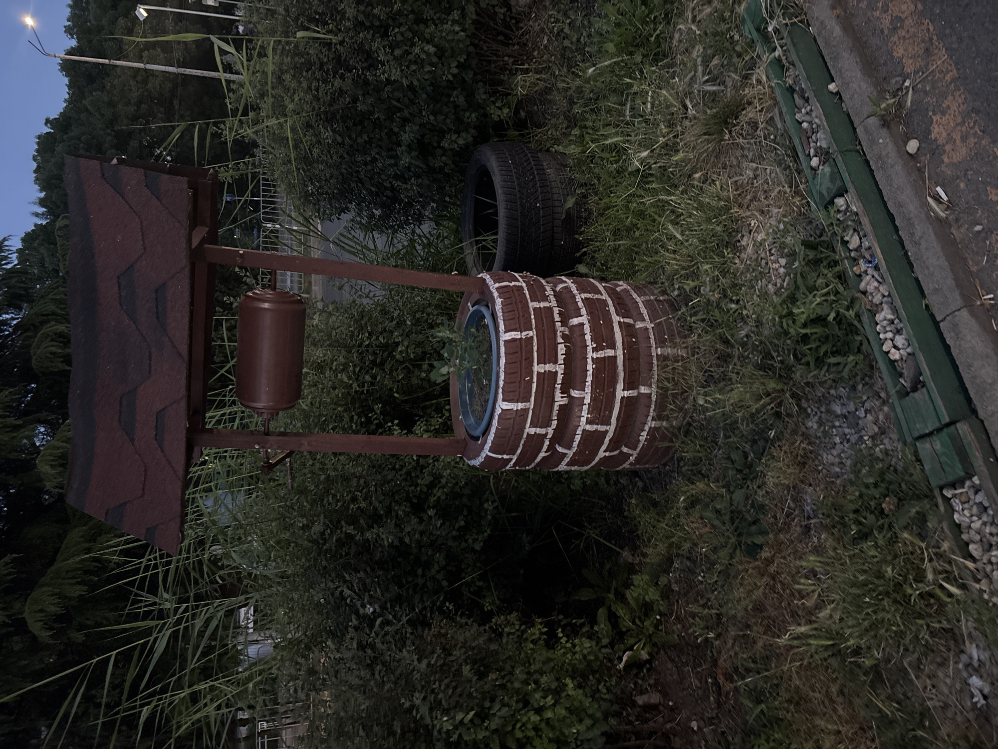 This image depicts a decorative well structure made from stacked tires painted to resemble bricks, with white mortar lines. The well has a red, shingled roof and a brown cylindrical water barrel or mechanism attached to a wooden support. It is situated outdoors, surrounded by lush green vegetation, including tall reeds and bushes. A second, unpainted black tire is visible next to the well. In the background, trees line a street, and a lamppost with a light on is visible. The ground is covered with grass and gravel, and a dark asphalt surface with a green wooden border runs along the foreground. The scene appears to be in a public or semi-public outdoor space, possibly a park or roadside, in Burgas, Bulgaria. There are no people or animals present in the image. The lighting suggests it is dusk or early evening.