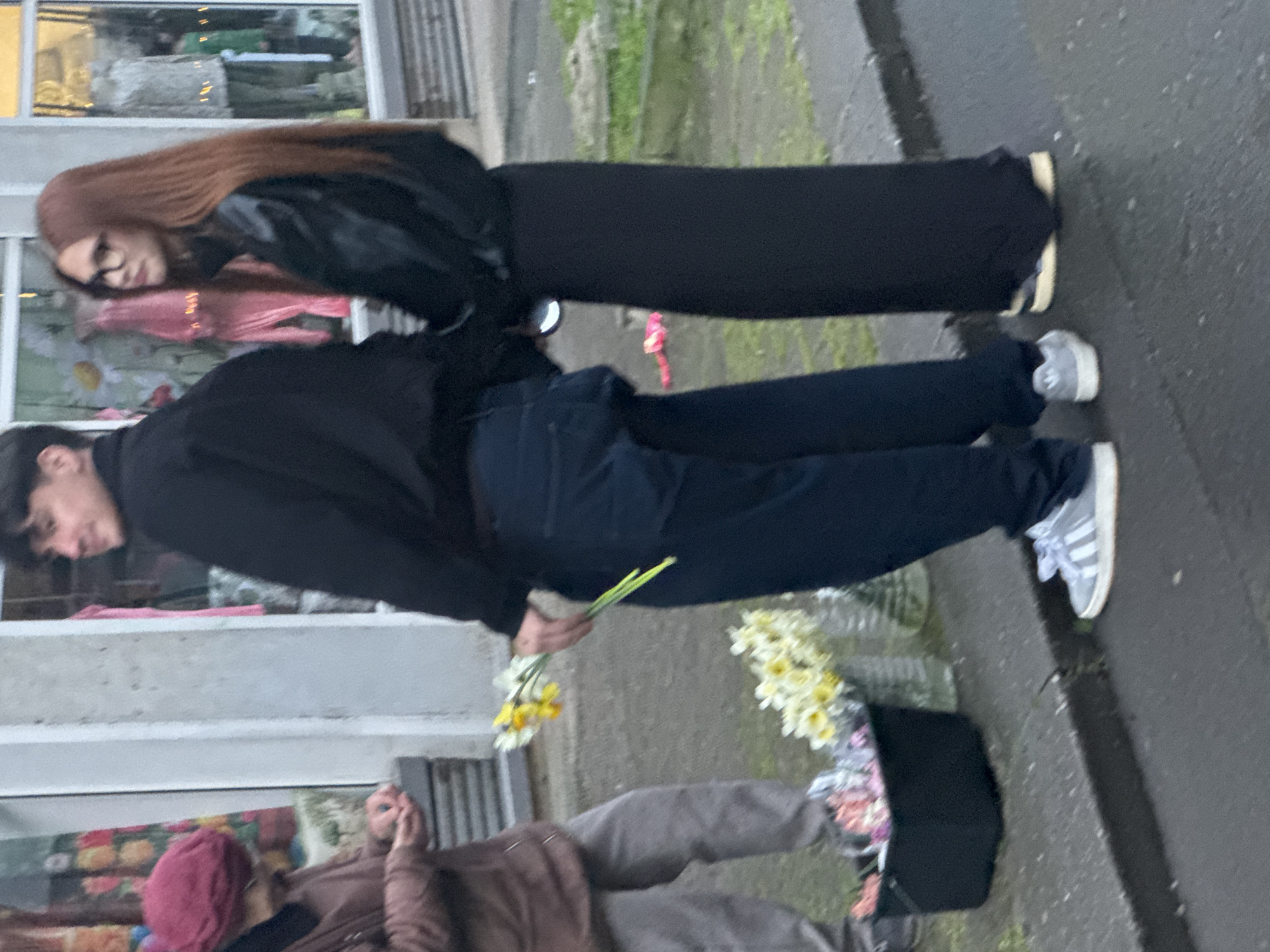 Two individuals, a male and a female, are standing on a city sidewalk. The male is holding a small bunch of daffodils. Another person is present in the foreground, and is looking at a cellphone. A flower shop is visible in the background. The exact location in Burgas, Bulgaria, cannot be determined from the image.