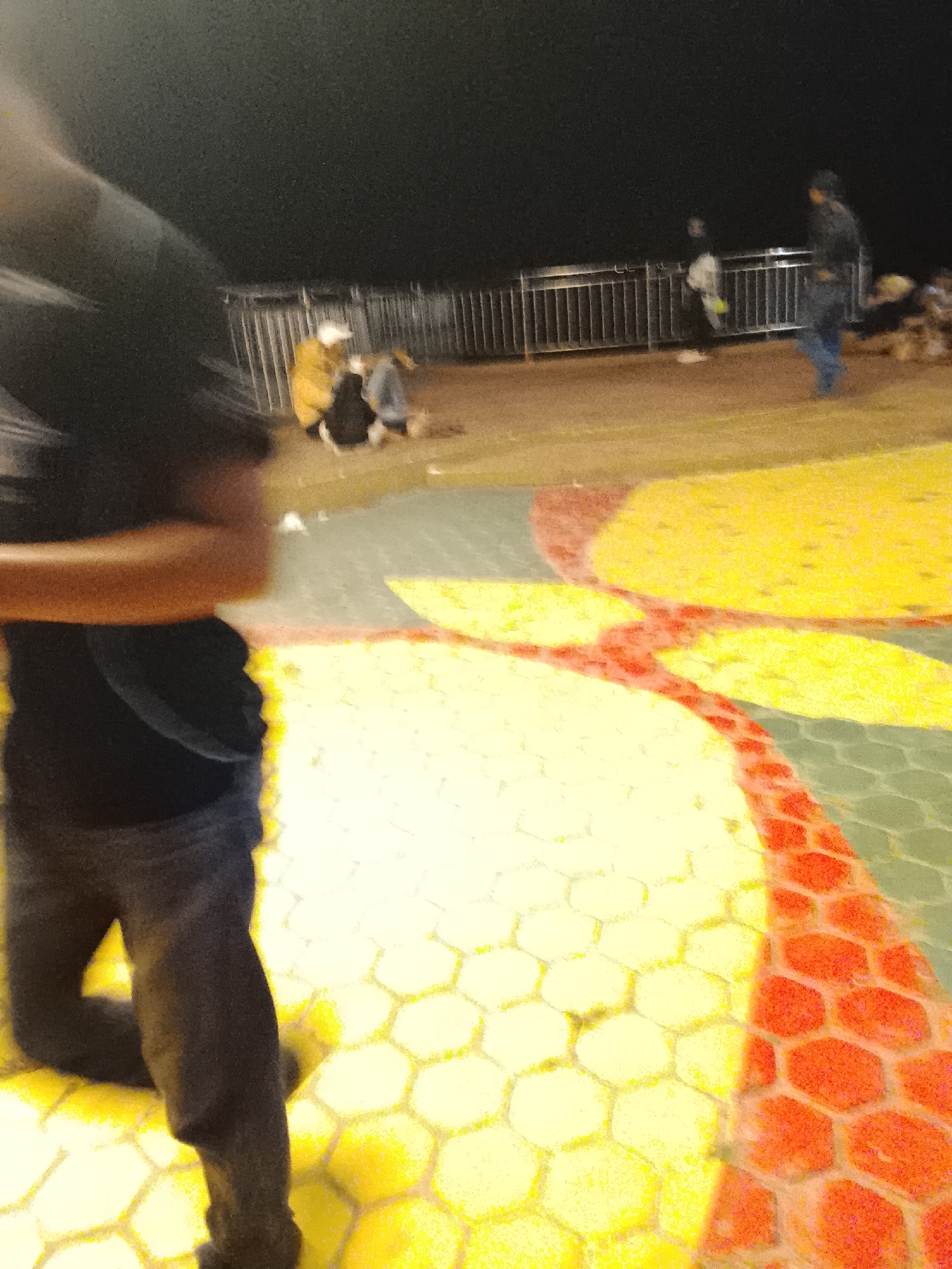 The image captures an outdoor scene at night, possibly a public space with a decorative ground pattern. In the foreground, a person is visible from the waist down, wearing dark jeans and a black t-shirt. The ground features a honeycomb-like pattern in yellow and red hexagonal tiles. In the middle ground, several people are gathered near a metal railing. One individual is wearing a yellow jacket. In the background, the scene is darker with indistinct figures and structures. The overall image quality is low, with motion blur affecting details. The provided context suggests the location is the city of Tanjung Balai Karimun, Indonesia, likely a waterfront promenade or public square due to the railing and open space.