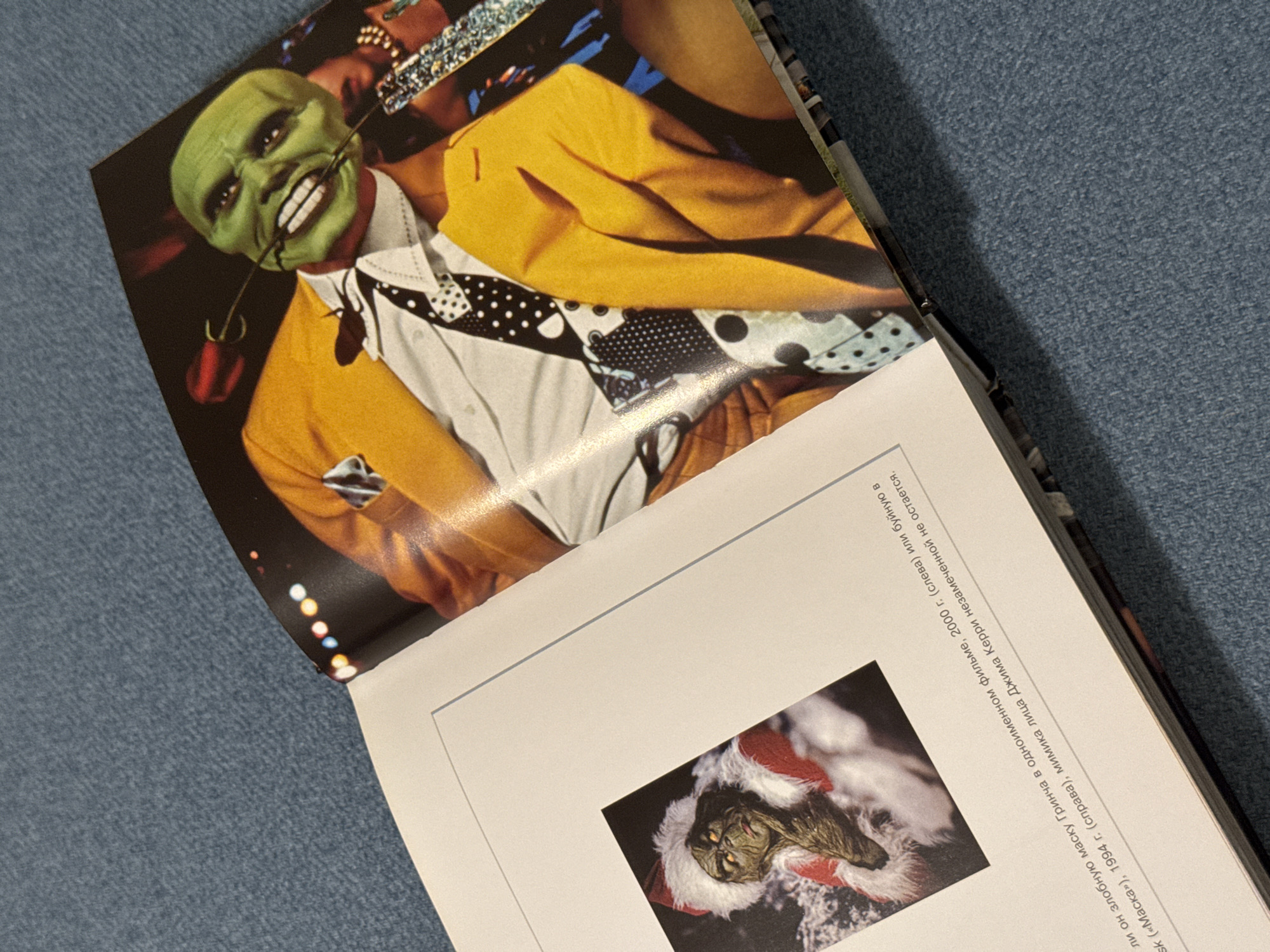 The image shows an open book. The left page features a picture of "The Mask" character, with a green face, yellow suit, and a rose in his teeth. The right page contains a picture of "The Grinch" character. The text near "The Grinch" image mentions the years 2000 and 1994, and Jim Carrey. The background of the image is a textured, blue surface. It is not possible to determine if this image was taken in Izhevsk, Russia.
