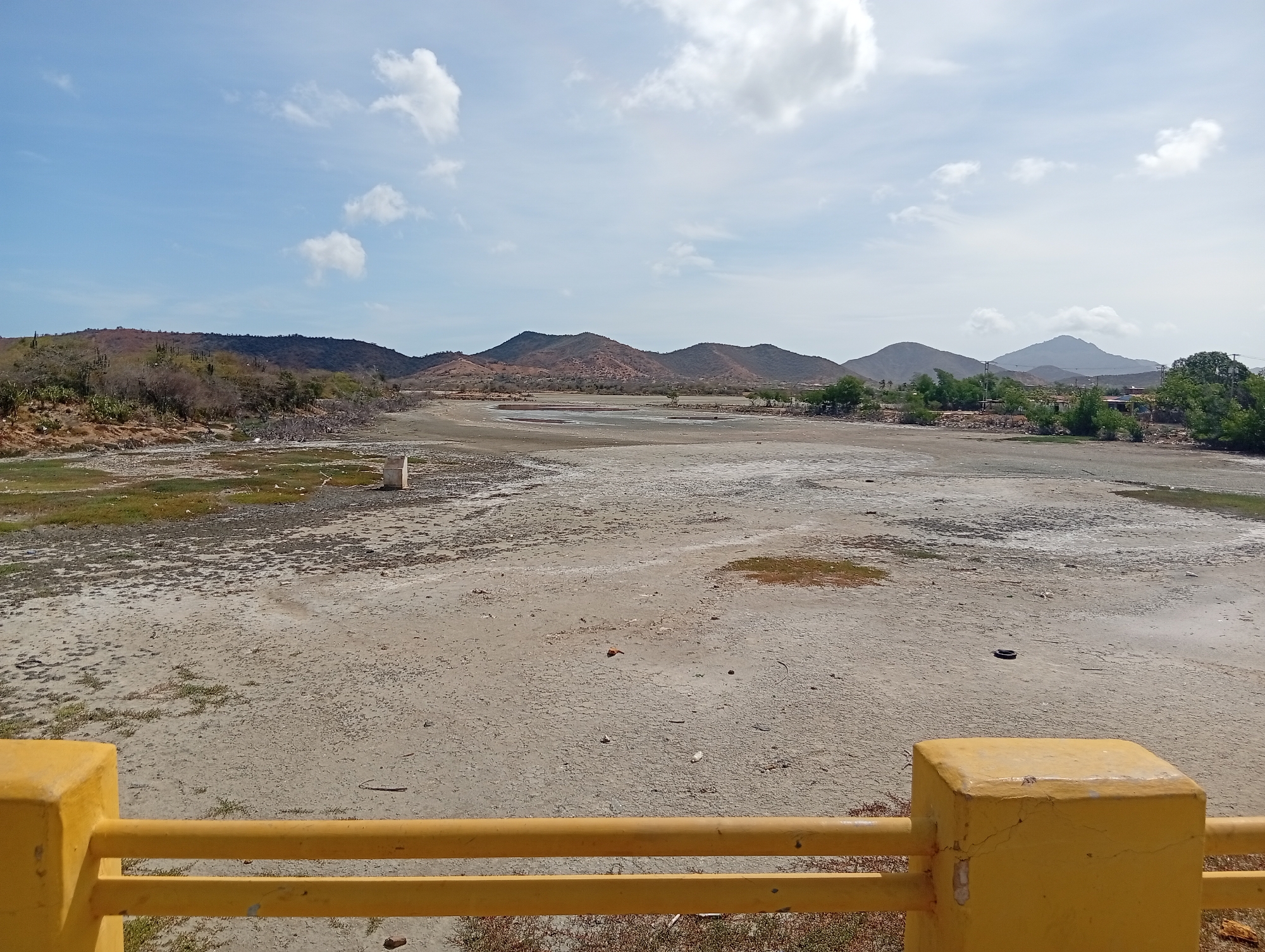 The image shows a dry, cracked landscape in Juan Griego, Venezuela, with a yellow bridge railing in the foreground. In the distance are rolling hills and mountains under a partly cloudy sky. The landscape is mostly barren, with some sparse vegetation. There's a tire on the dry land and some debris scattered around. The overall impression is of aridity and desiccation.