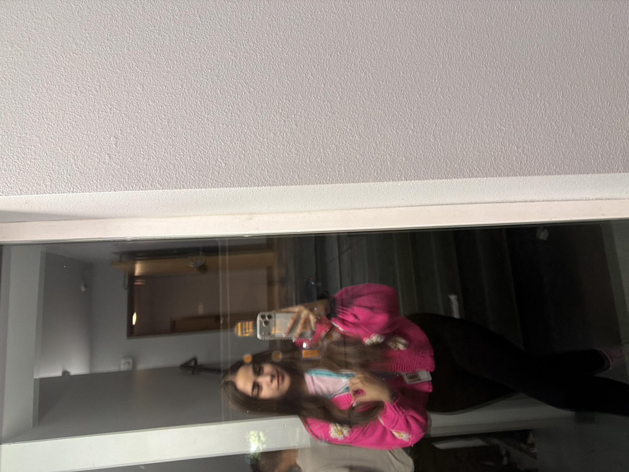 A young woman is reflected in a floor-to-ceiling mirror. She is wearing a bright pink cardigan with floral embellishments over a pink t-shirt and black leggings. She is holding a smartphone in her right hand, taking a selfie. A lanyard with an ID card is visible around her neck. The background of the reflection shows a hallway with doors and stairs. The environment appears to be indoors, possibly a building in Lake Delton, Wisconsin, United States of America. The wall to the right of the mirror is textured and light-colored.