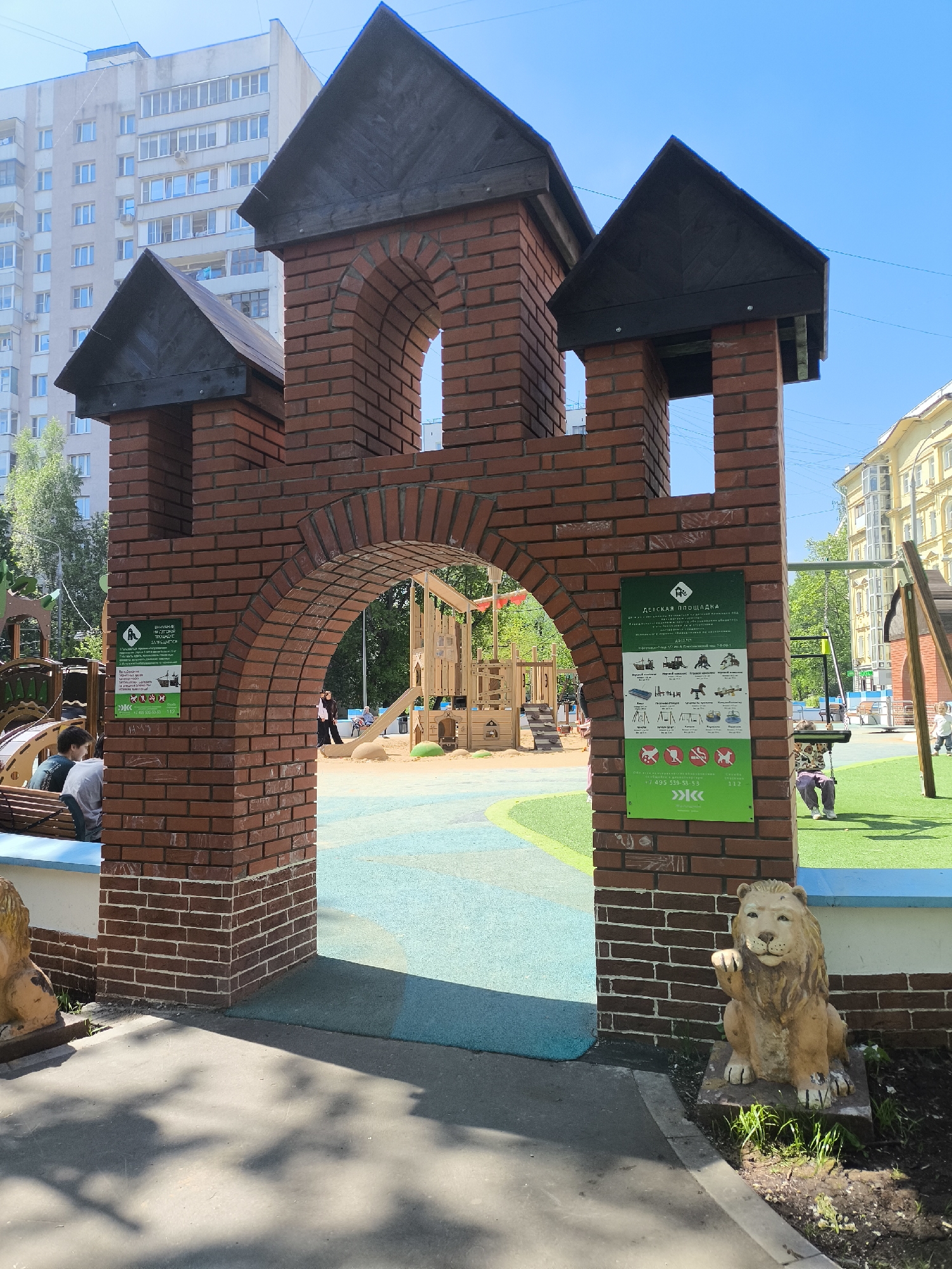 The image shows a playground entrance in a city. The entrance is a brick structure resembling a castle gate, with two towers topped by black, gabled roofs. A brick arch forms the opening. Each side of the entrance has a green sign with text in Russian. At the base of the right side, there is a lion statue.
The playground inside the gate features a sand area and various wooden play structures. Artificial grass covers part of the playground surface. Some people are visible within the playground, including children playing on the equipment.
Outside the entrance, there is a concrete pavement with tree shadows cast upon it. In the background, there are several apartment buildings and trees.