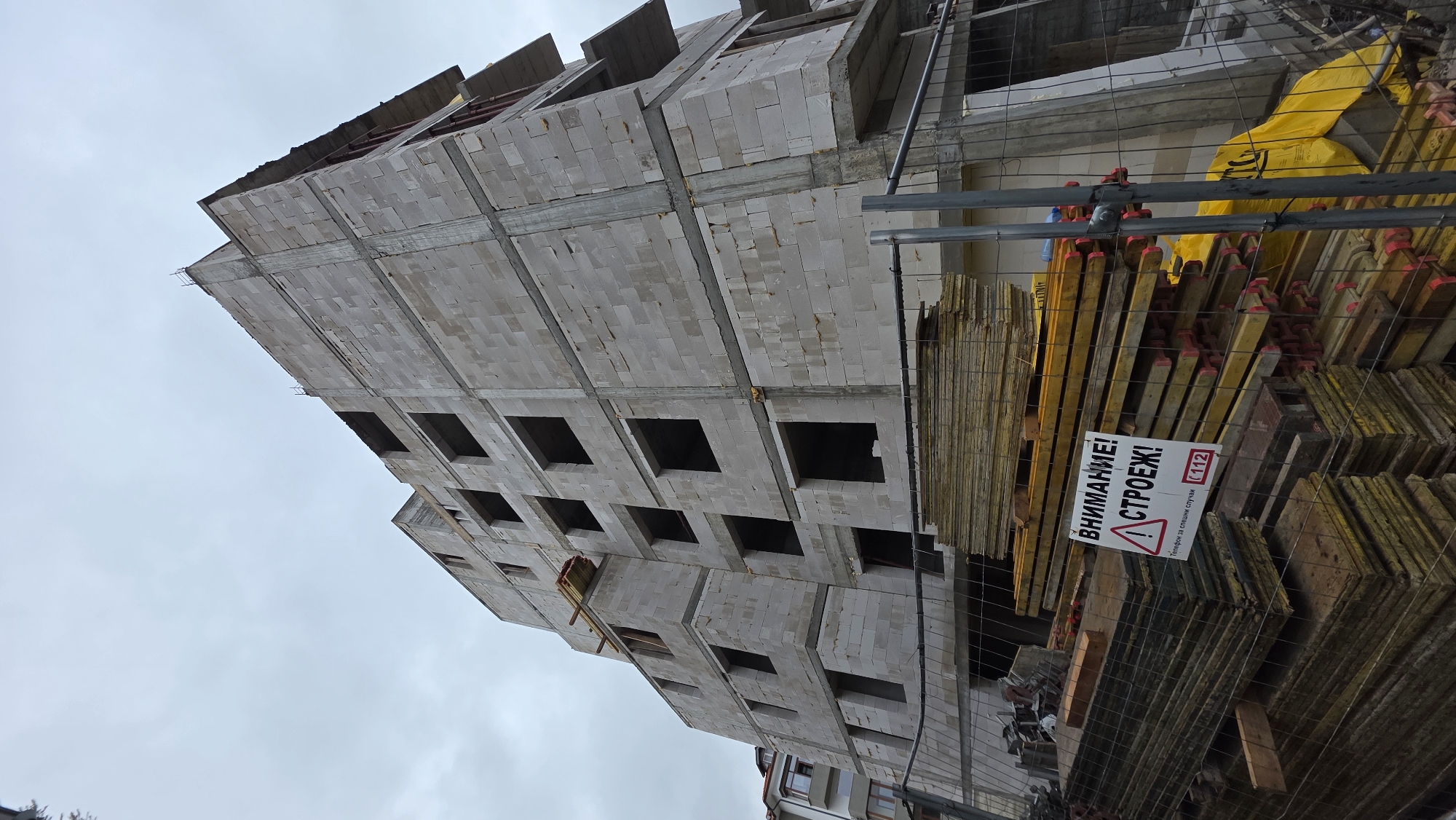 The image shows an unfinished multi-story building under construction in Blagoevgrad, Bulgaria. The building is made of white blocks with regularly spaced window openings. Construction materials, including yellow planks, are stacked behind a chain-link fence in the foreground. A sign in Cyrillic script warns of the construction site and provides an emergency contact number. The sky is overcast.
