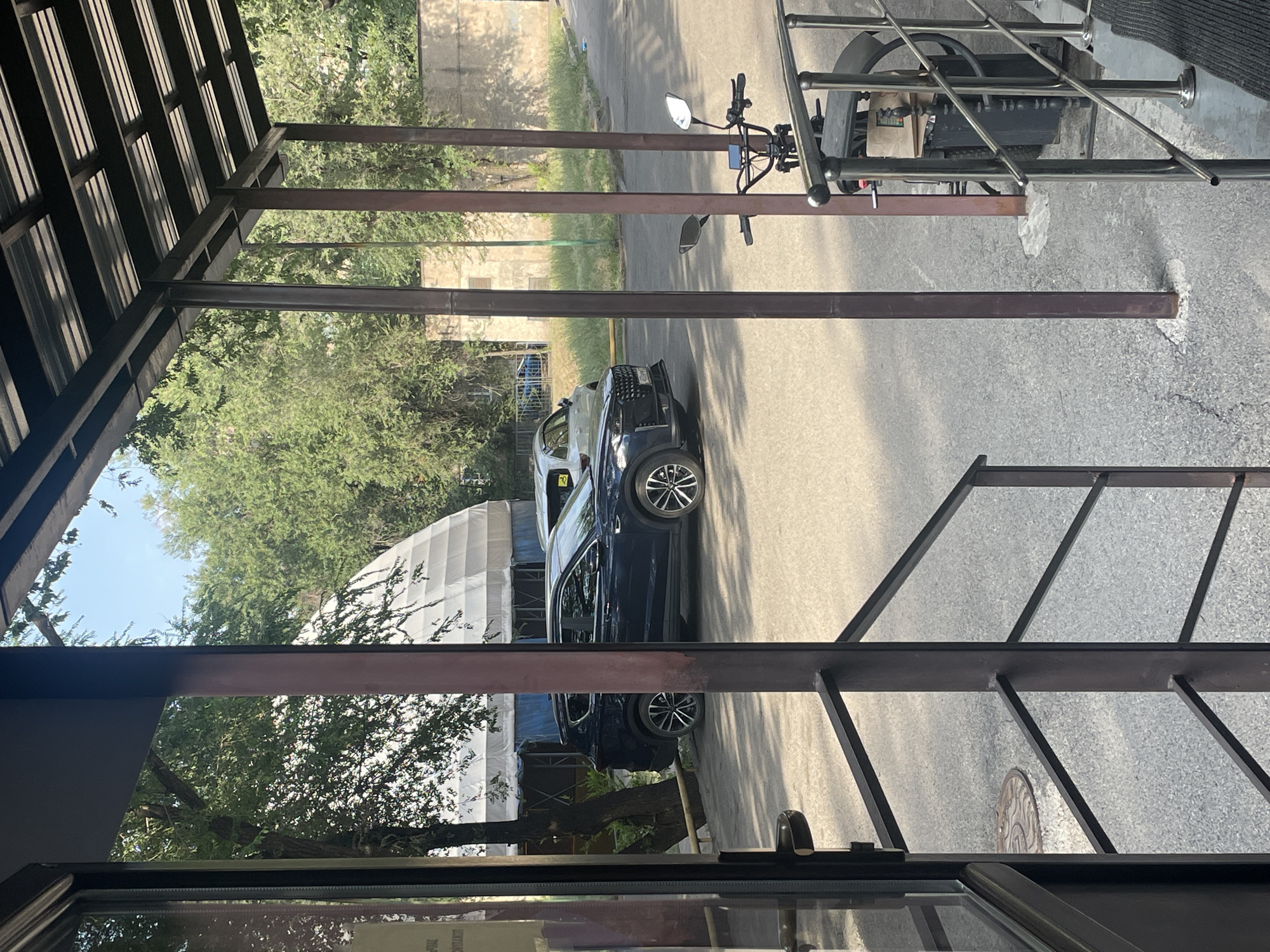The image depicts an outdoor scene in the city of Almaty, Kazakhstan, viewed from under a covered walkway. In the foreground, a dark blue SUV is parked on an asphalt surface. To the right of the SUV, a black electric scooter is parked. Beyond the vehicles, a white tent structure is visible amidst lush green trees. The overall environment suggests a residential or commercial area with ample greenery and clear skies. The lighting indicates it is daytime.