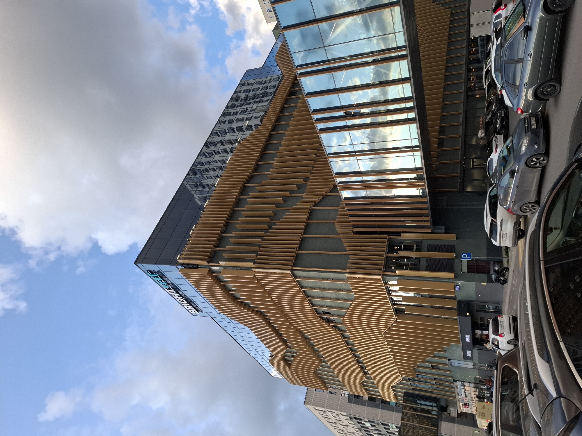 The image shows a modern building in Tyumen, Russia, with a distinctive facade of vertical golden louvers. The building appears to be a commercial or office complex, with the "BRIGHT FIT" logo visible on the upper portion. A pedestrian walkway with glass walls and golden framing connects different parts of the building. In the foreground, several cars are parked on a street in front of the building, suggesting an urban environment. The sky above is partly cloudy, with a mix of blue sky and gray clouds. The overall scene depicts an architectural landmark in a city setting.
