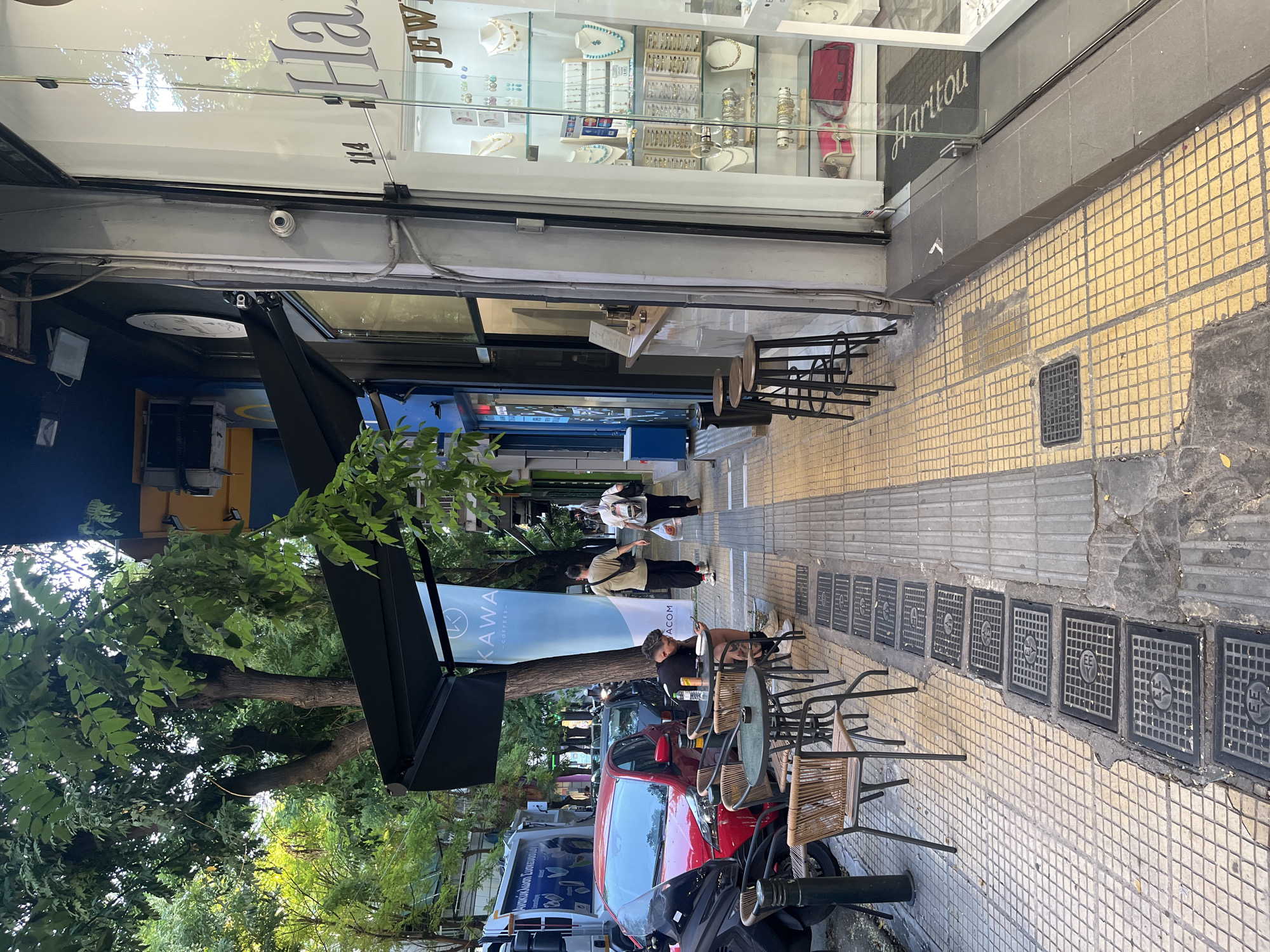 This is a street-level photograph taken in Athens, Greece, likely in a commercial area. The scene depicts a sidewalk with outdoor seating for a cafe. A man with tattoos is seated at a small table, looking at his phone. Two other individuals are walking away from the viewer down the sidewalk. To the right, a jewelry store's display window is visible, showcasing necklaces, earrings, and other accessories. A black awning extends from the building above the cafe seating. Trees line the street, providing shade. A red car and a motorcycle are parked along the curb. The sidewalk is paved with yellow and grey tiles and features several manhole covers. The overall impression is a bustling urban environment.