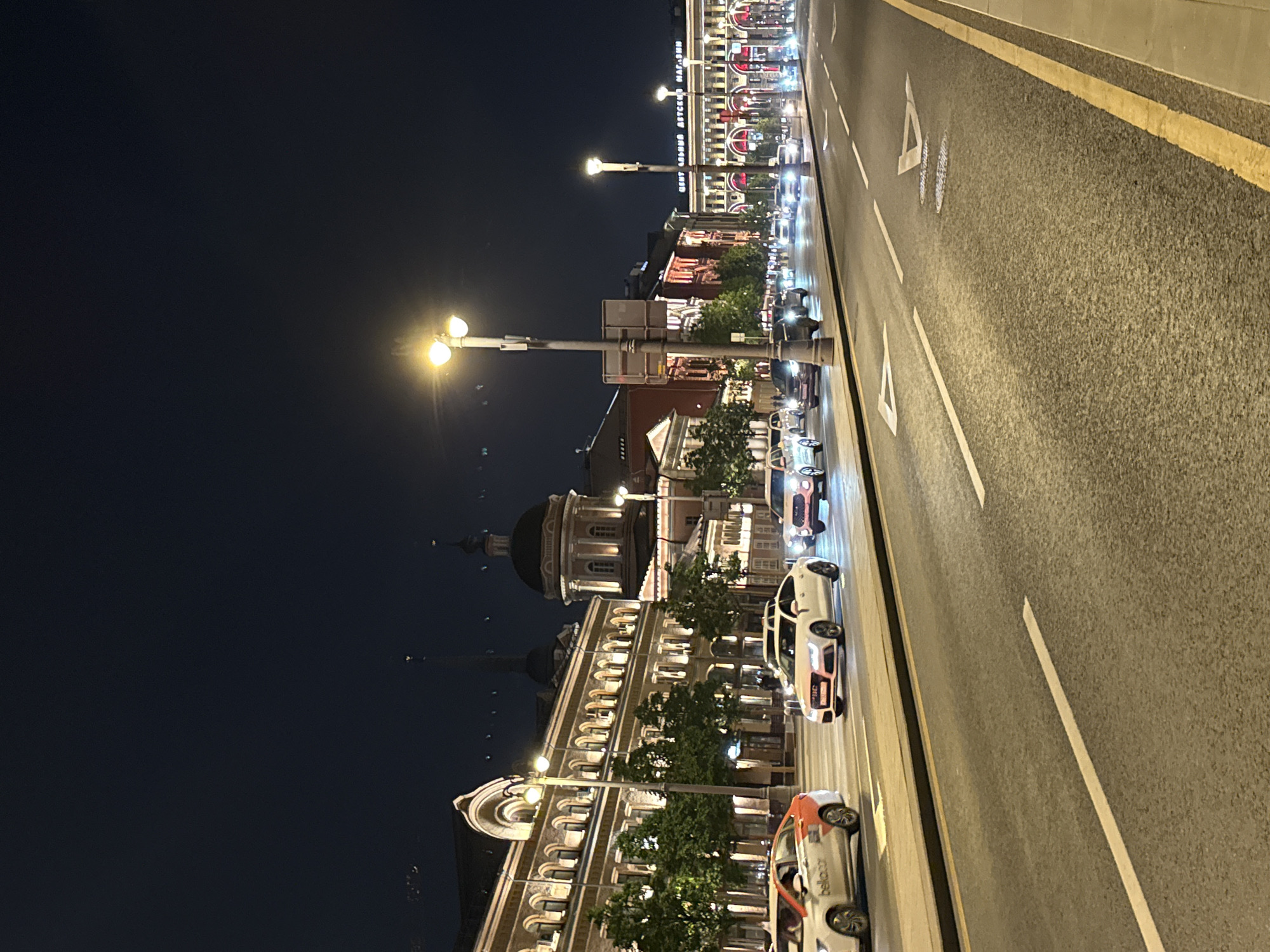 The image shows a nighttime street scene in Moscow, Russia. A wide, multi-lane road with yellow and white lines runs along the right side of the image. Buildings, illuminated with streetlights, line the left side of the road. Cars are traveling on the road. The architecture appears ornate, with domes and detailed facades. Streetlights are visible along the road. The sky is dark.