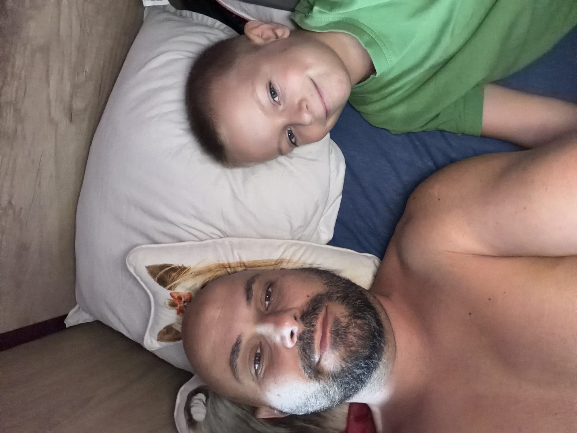 This media file features two individuals, a child and an adult male, lying down in a room. The child, wearing a green t-shirt, is positioned above the adult on a white pillow. The child has light skin and light brown hair, with bright eyes looking towards the camera. A gentle smile is visible on their face. The adult male, lying on another pillow with a decorative pattern, has a shaved head and a beard. His eyes are open and he is looking directly at the camera. He appears shirtless, revealing his chest and shoulder. The background shows a blue surface, possibly a bed or couch, and a wooden element, suggesting a room setting. The lighting indicates it is daytime. The overall mood is relaxed and intimate, possibly during a moment of rest or play between the two individuals. No visible text is present in the image. The location context provided is Dolni Dabnik, Bulgaria.