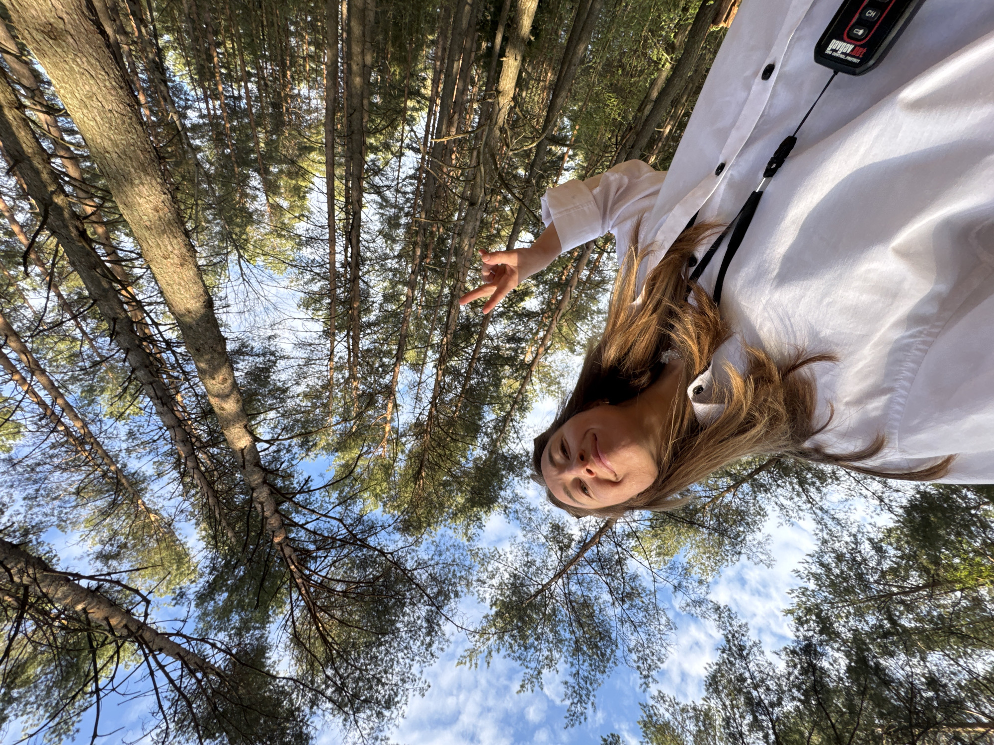 A woman is in a forest in Прикамский, Russia, looking up at the camera. The woman is wearing a white shirt with a black lanyard holding a Geiger counter attached. Her hair is brown. The photo has been taken from a low angle looking upwards, showcasing the tall trees overhead. The sky is visible through the trees and is light blue with white clouds.