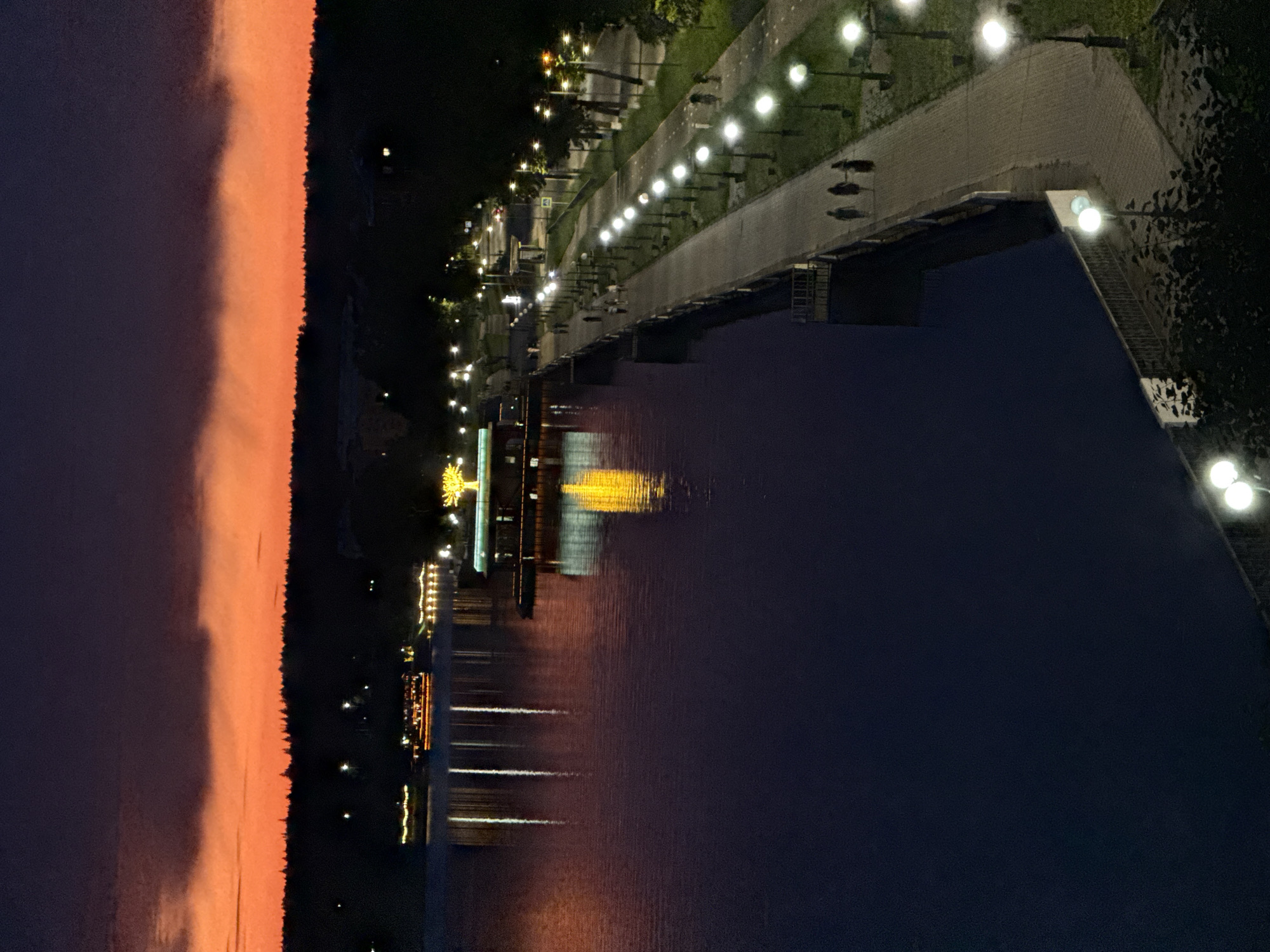 This is an aerial, nighttime photograph of the Votkinsk Reservoir embankment in Votkinsk, Russia. The sky displays a gradient of dark purple to vibrant orange from top to bottom, indicating either dusk or dawn with a cloud cover. The reservoir, on the left side of the image, is dark blue with reflections of lights from boats and structures along the opposite shore and embankment.
On the far left, several boats are illuminated with strings of yellow lights, their reflections extending vertically into the water. In the mid-ground, a pier extends into the reservoir. On this pier, a structure is visible, illuminated by bright yellow lights that create a tree-like shape, with its reflection prominently displayed in the water.
The embankment, on the right side of the image, is lined with a paved walkway and numerous streetlights, each casting a bright white glow. The walkway curves away from the reservoir and disappears into the background. Several trees are scattered along the embankment.
A few individuals are walking along the embankment. In the foreground of the embankment, three figures are walking away from the camera. Further up the embankment, several other figures are walking in both directions. The scene depicts a peaceful evening or morning ambiance on the Votkinsk Reservoir embankment.