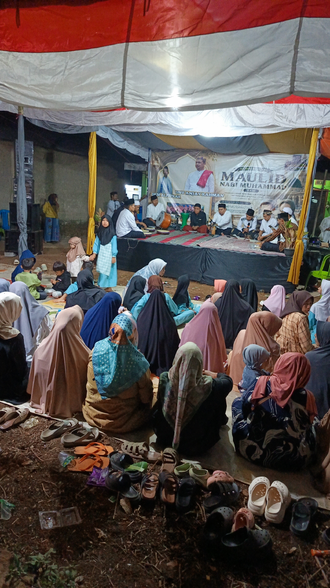 This image depicts an outdoor gathering in Margajaya, Indonesia, likely for a religious event given the banner which reads "MAULID NABI MUHAMMAD." A group of people, predominantly women and children, are seated on the ground in the foreground, many wearing hijabs of various colors and patterns. Some are looking towards a raised stage area in the background, where a group of men are seated. The stage is covered by a tent-like structure with a red and white striped cloth at the top. On the stage, there's a large banner featuring an image of a man and text in Arabic and Indonesian. Some men on the stage appear to be playing musical instruments, possibly for a performance or recitation. The ground is uneven and appears to be dirt, with numerous pairs of sandals and shoes scattered around the seated attendees, indicating they have removed them for the event. The overall atmosphere seems to be one of communal gathering and religious observance.