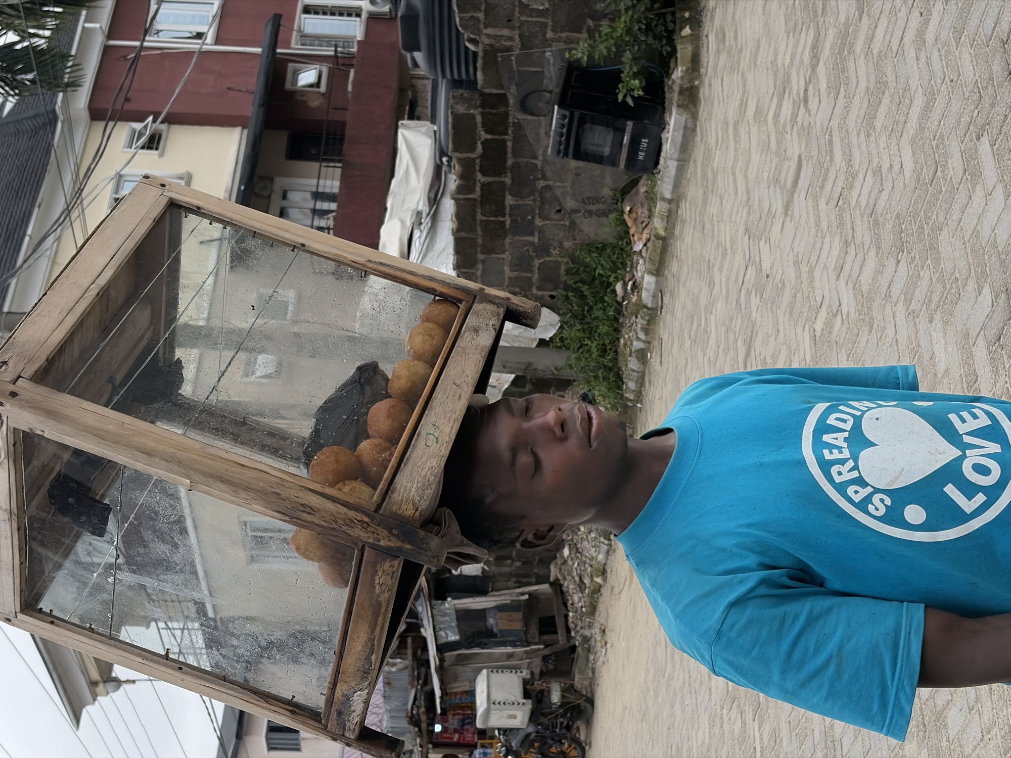 A young male, wearing a teal t-shirt with "SPREADING LOVE" printed on it in white, walks outdoors carrying a wooden and glass display case filled with round, golden-brown snacks balanced on his head. The display case is tilted forward, with its contents visible through the glass panels. In the background, a multi-story building with a red and yellow facade is partially visible, along with other structures and debris that suggest an urban setting. Power lines crisscross overhead. The ground is paved with interlocking bricks, and a brick wall with various objects, including what appears to be a black oven, is visible to the right. The scene is set in Igboefon, Nigeria.