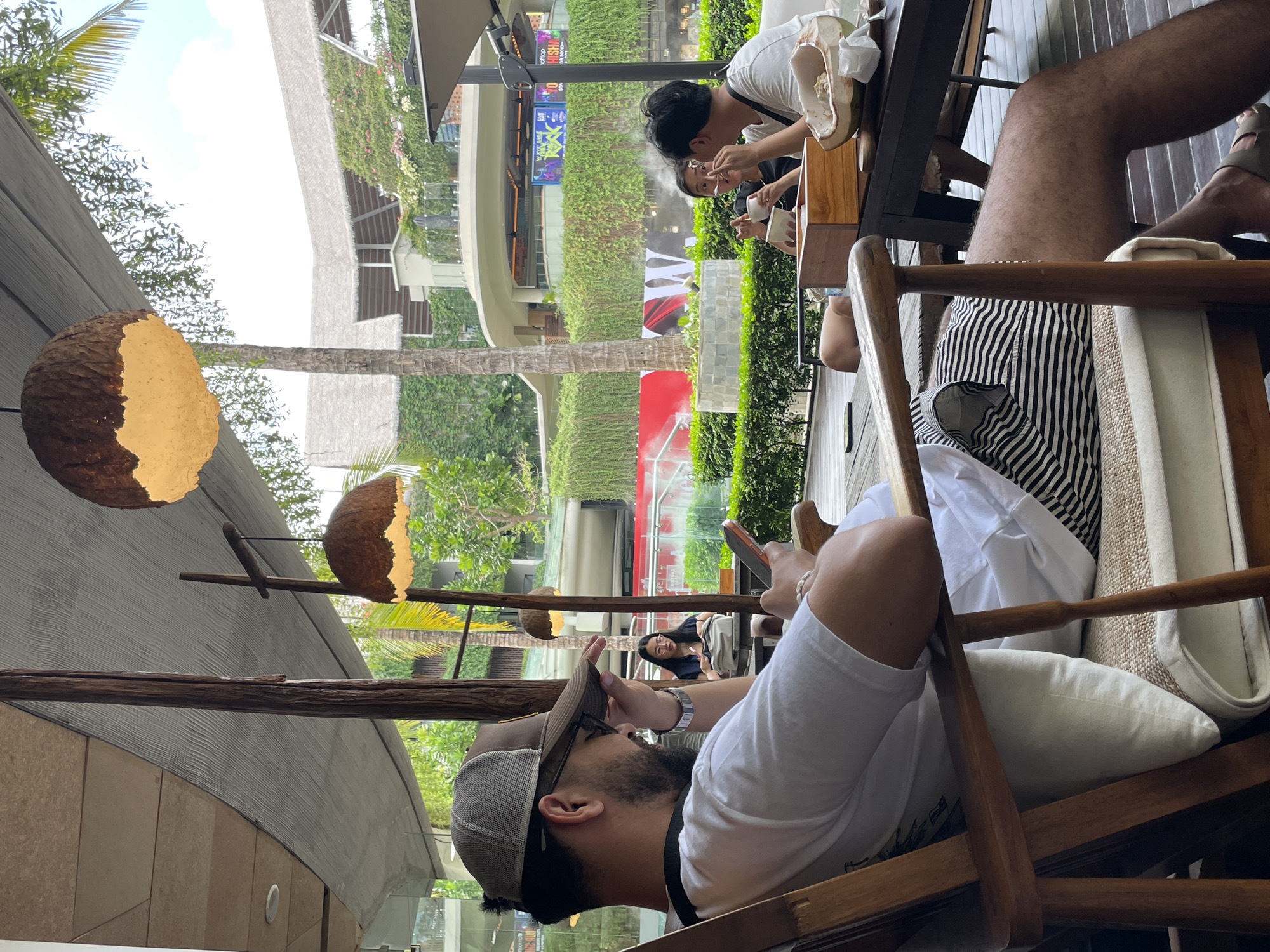 This image depicts an outdoor seating area in Kuta, Indonesia, likely at a restaurant or cafe, with lush greenery and a multi-story building with a curved facade in the background. In the foreground, a man wearing a baseball cap, glasses, and a white t-shirt is seated in a wooden chair, looking at his phone. He has a beard and is wearing striped shorts. To his right, at a wooden table, two individuals are seated. One, a woman with glasses and dark hair, is holding a utensil and looking at her food. Another person, partially visible, is also seated at the table. On the table, there is a halved coconut with a spoon and some food. The environment features natural lighting, with overhead coconut shell pendant lamps casting a warm glow. Wooden beams and a textured canopy provide shade over the seating area. The background reveals a building with extensive vertical gardens and an elevated walkway.