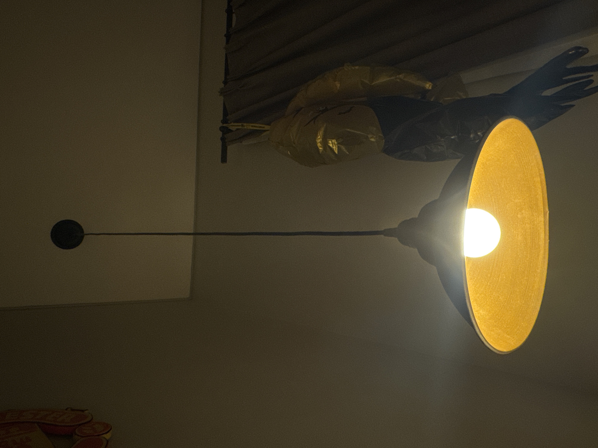 The image shows a dimly lit interior in Kuta Selatan, Indonesia. An industrial-style lamp with a large, yellow, bowl-shaped shade is illuminated, casting a bright light. Above the lamp, some metallic gold balloons and black streamers are attached to what appears to be a window blind fixture. A thin, dark rod extends horizontally from the lamp towards the left, ending in a small, dark circular fixture. At the bottom-left corner, parts of a stack of red and yellow objects are visible. The walls are beige and out of focus. The overall environment appears to be a room with low lighting.