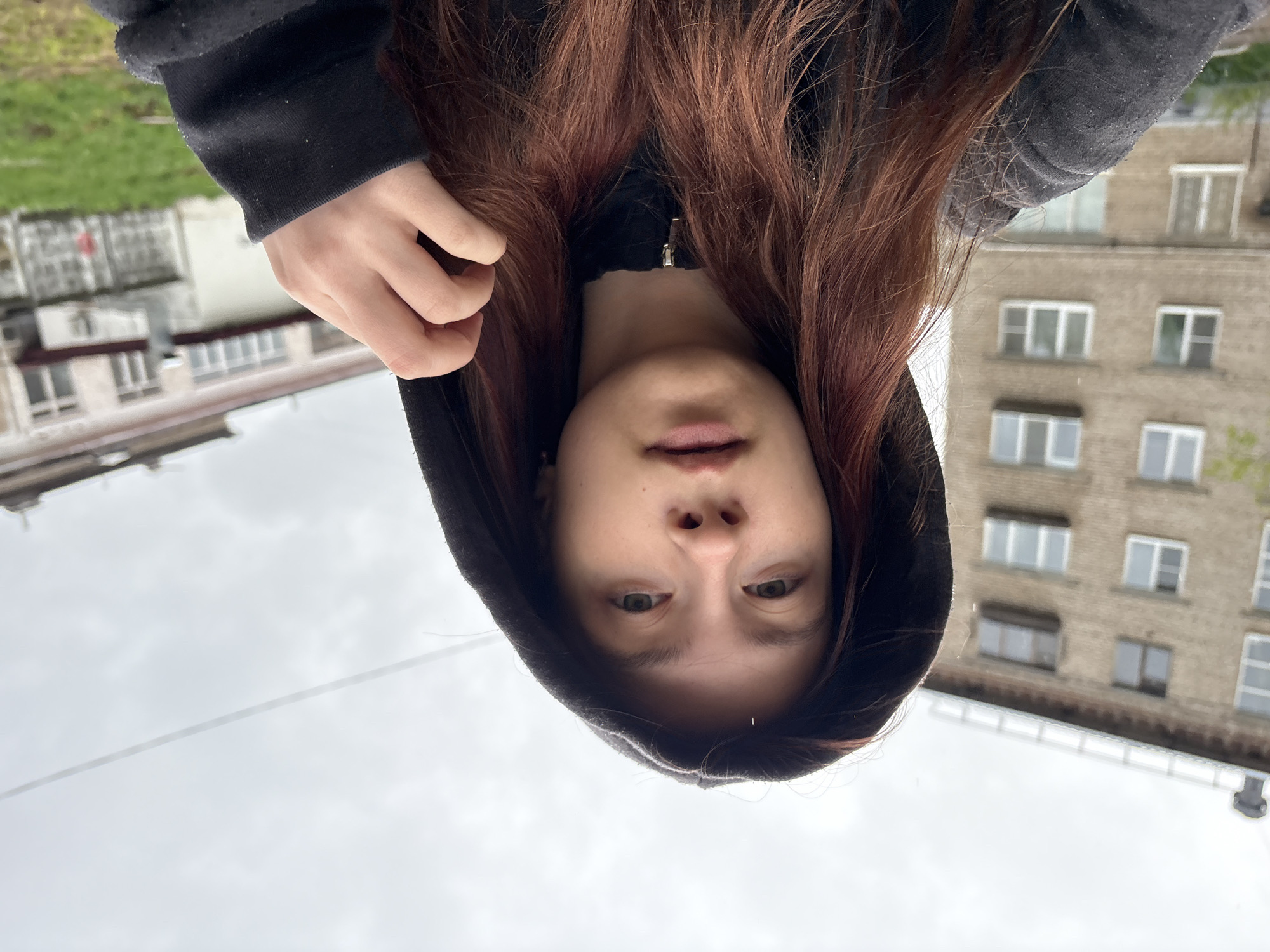 A person wearing a black hooded sweatshirt is holding up a smartphone to take a selfie. The person has long, reddish-brown hair. The background shows a multi-story apartment building constructed of brick. Other buildings and hints of greenery are visible in the distance. The photograph appears to be taken outside in overcast weather in Chaikovsky, Russia.