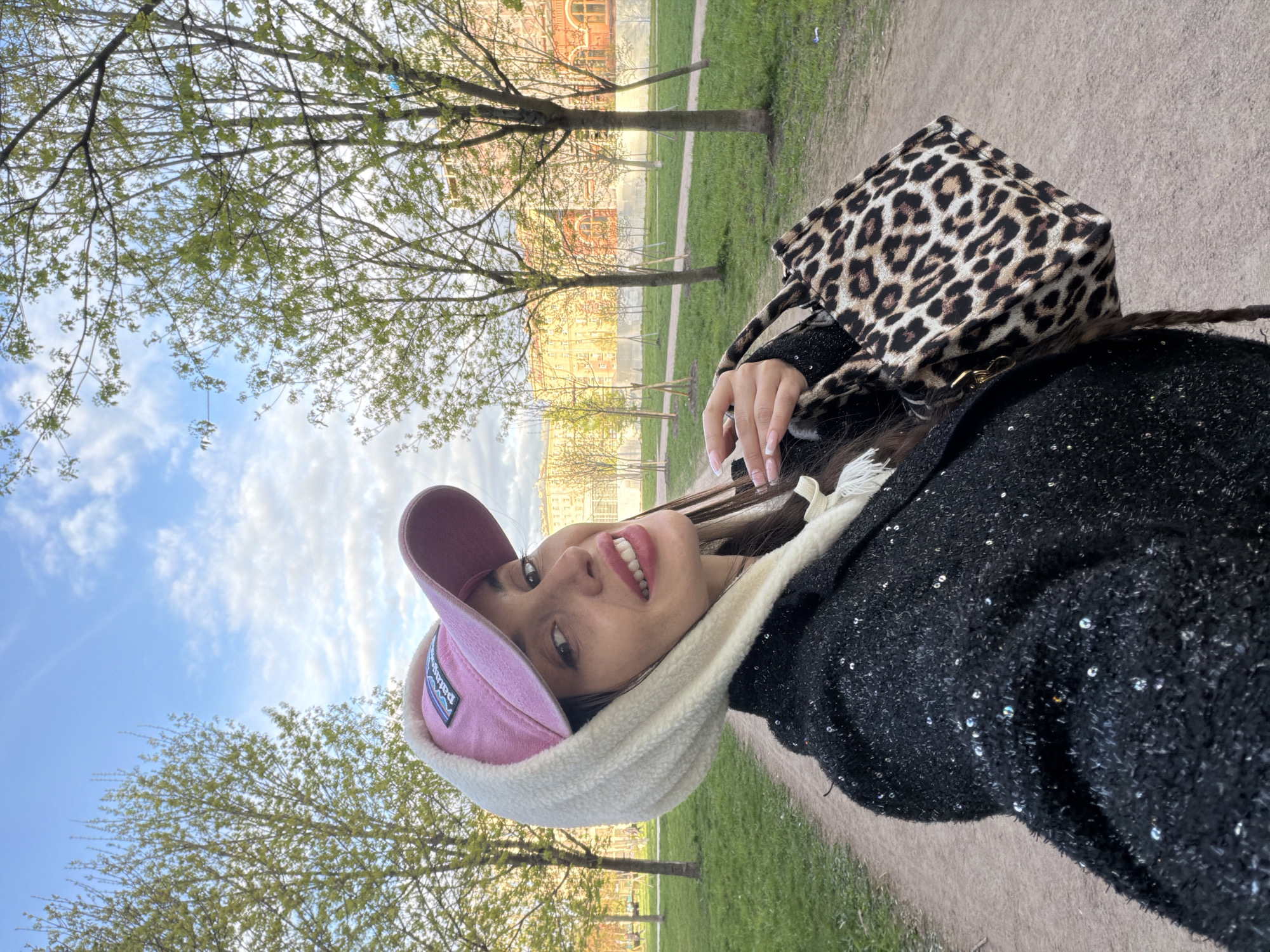A selfie taken in Saint Petersburg, Russia, shows a young woman wearing a pink baseball cap, a white scarf, and a black, sequined jacket. She also has a leopard print handbag. The background consists of trees, grass, a path, and a building in the distance. The sky is blue with scattered clouds.