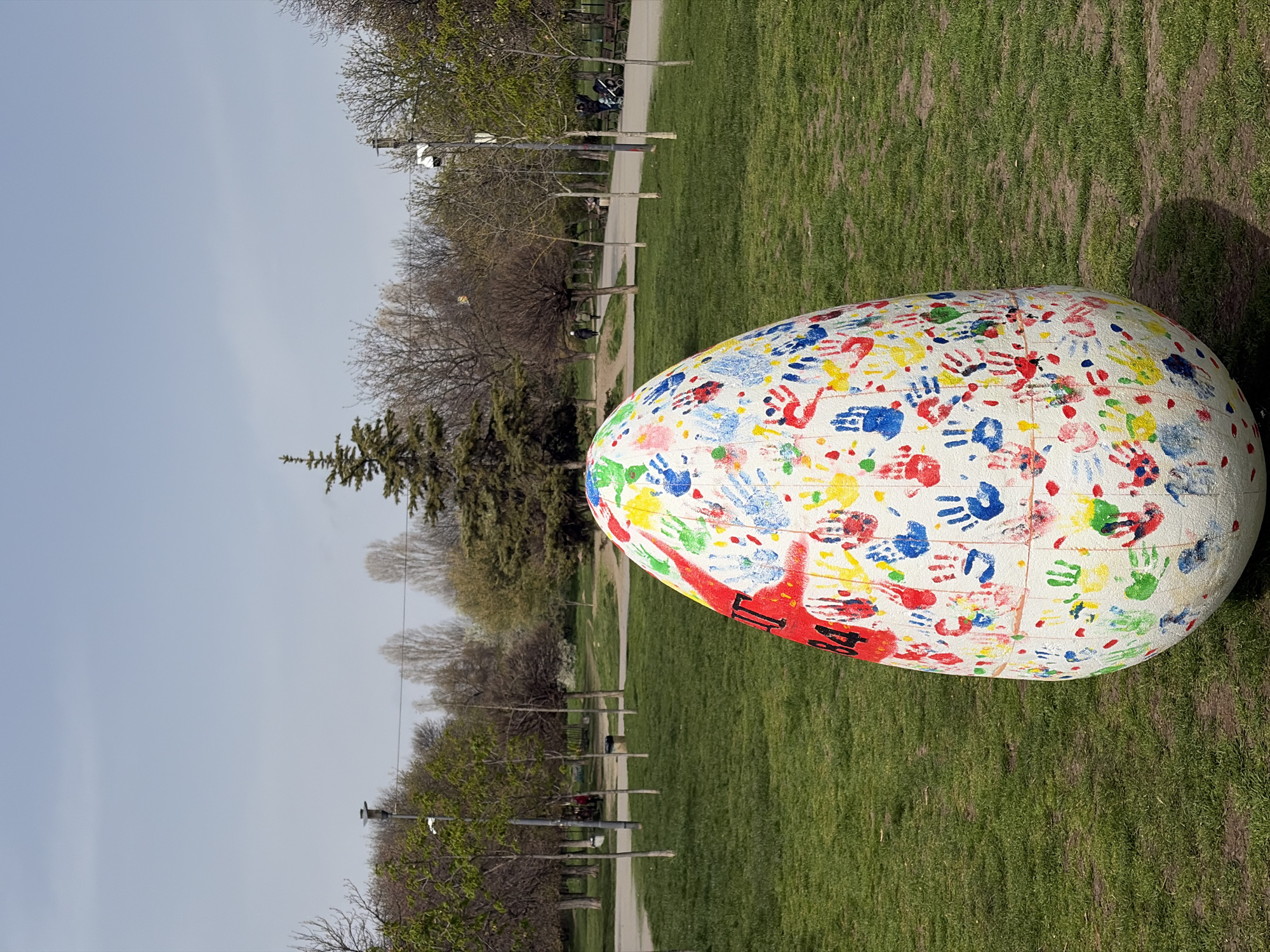 The image shows a large, white Easter egg sculpture covered in colorful handprints, sitting on green grass in a park. In the background, there are trees, walking paths, benches, and streetlights. The sky is overcast. The specific location is not discernible from the image, but it's said to be in Sofia, Bulgaria.