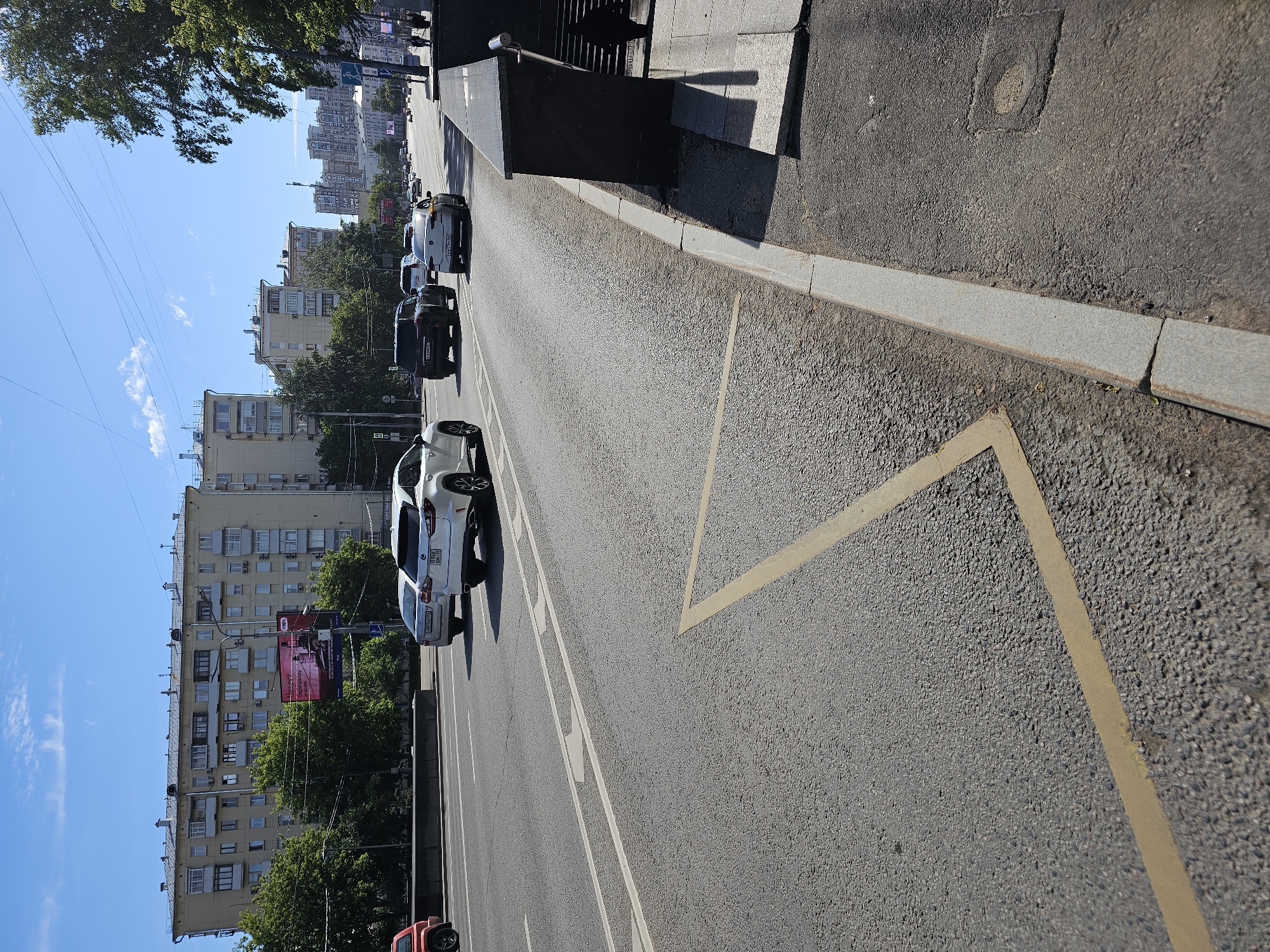 The image depicts a street scene in Moscow, Russia, under a clear blue sky with scattered clouds. The foreground features an asphalt road with white lane markings, including a yellow triangular warning marking. A grey concrete curb runs along the right side of the road, adjacent to a dark granite structure with a metal railing leading to stairs.
Several vehicles are present on the road. A white Lexus SUV is in the right lane, followed by a dark SUV. Further ahead, a white sedan is visible. Other vehicles are also seen in the distance.
On the left side of the road, there is a barrier and trees. Behind the trees, apartment buildings with multiple stories and visible windows are prominent. A large billboard with a pink advertisement is also visible on one of the buildings. Power lines stretch across the sky.
In the background, more buildings, some with distinct architectural features, and trees line the street, suggesting an urban environment within Moscow. The overall scene captures a typical day on a busy city street.