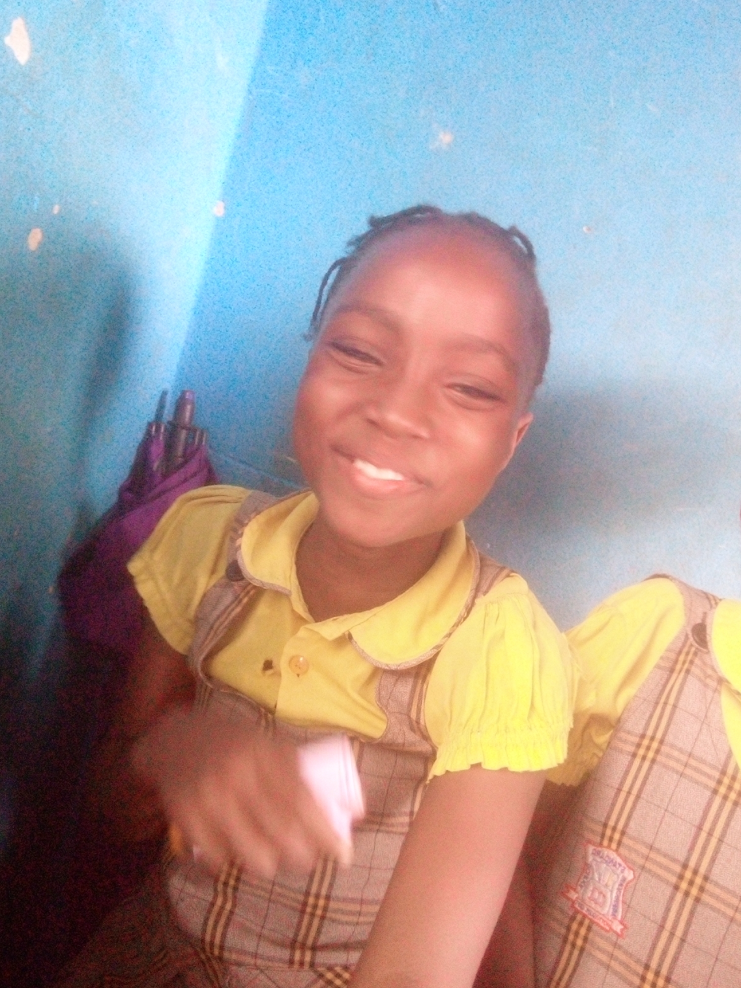 A young girl is photographed indoors against a light blue wall. She is smiling and wearing a yellow shirt with a ruffled collar and sleeves, paired with a plaid overall dress that has a school crest or emblem stitched on it. An umbrella is partially visible in the background on the left. A portion of another child wearing a similar outfit is visible on the right. The location is stated as Ikorodu, Nigeria.
