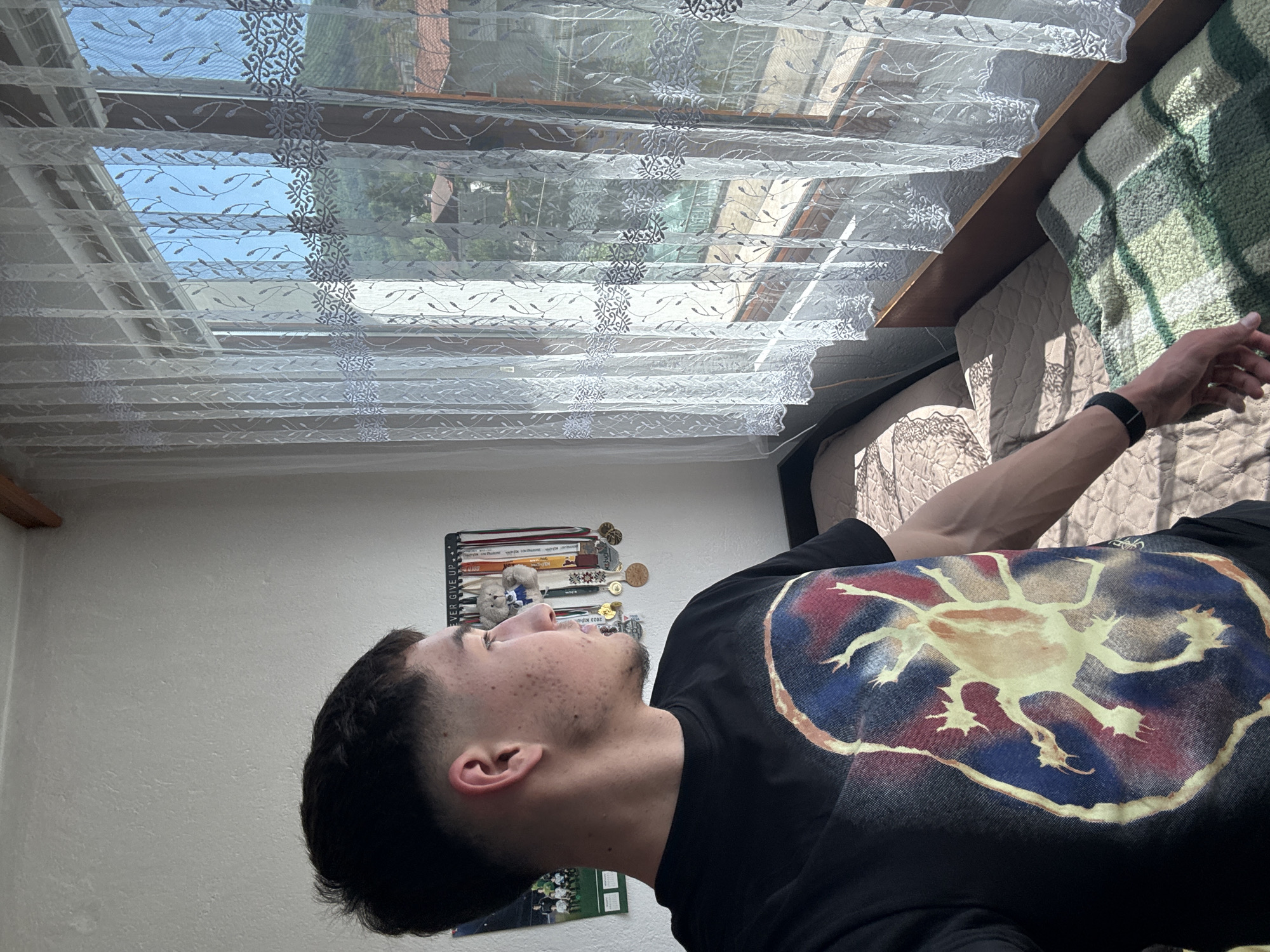 A medium shot depicts a young man with dark hair and light skin lying on a bed in an indoor setting. The man is wearing a black t-shirt featuring a graphic print of a stylized creature with multiple limbs or appendages against a background of blue, red, and gold. He is wearing a black fitness tracker on his left wrist and his right arm is extended with his hand visible.
A window is located in the top portion of the frame, partially covered by a patterned white curtain. The outside view through the window includes foliage and glimpses of buildings. The setting appears to be a residential building, and while the city of Zlatograd, Bulgaria is mentioned, there are no specific landmarks or indicators within the image to conclusively verify the location. A display of medals and other items is affixed to the wall, along with what appears to be a poster or print. The bed features a patterned blanket or bedding.