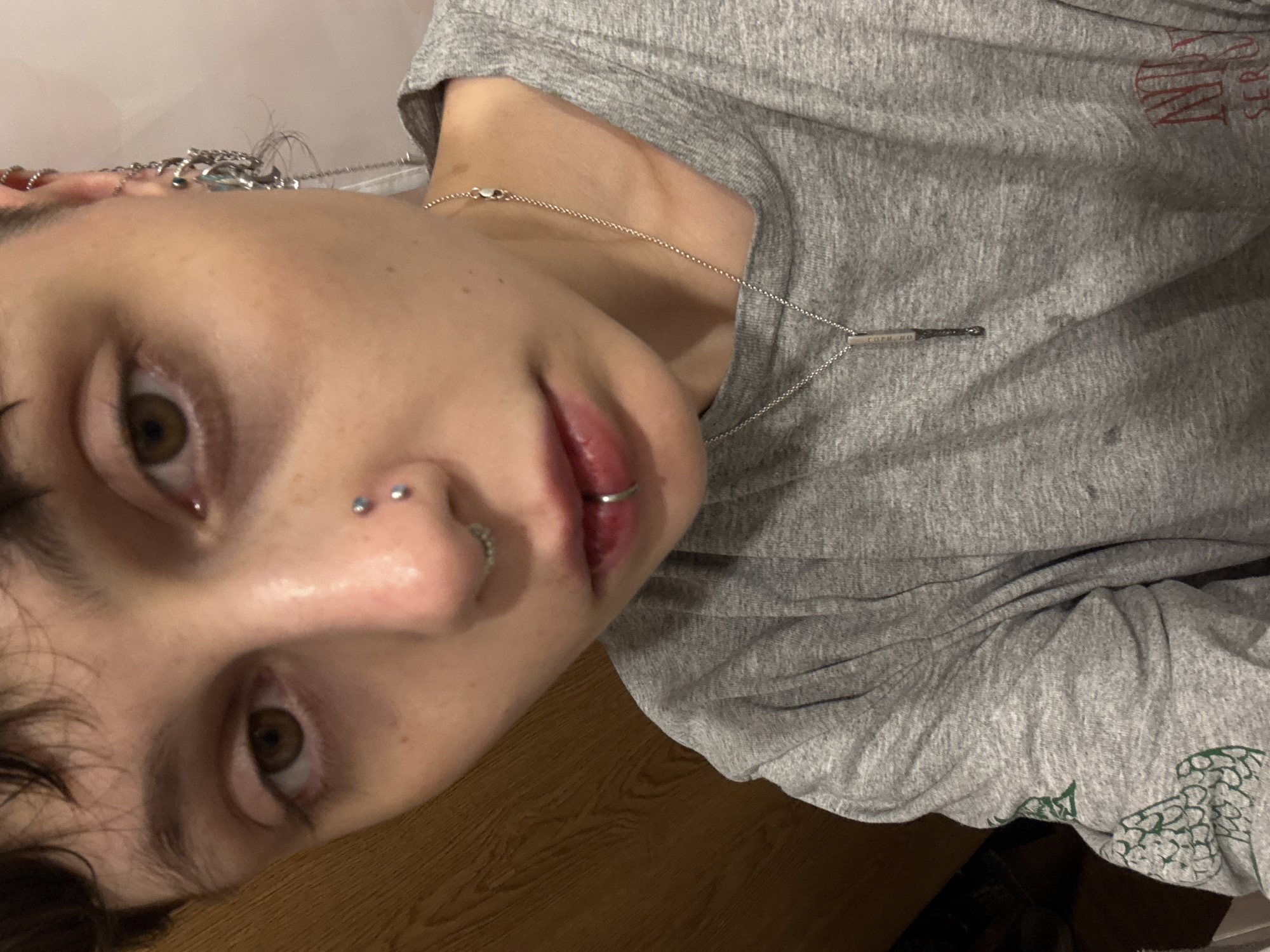 A close-up, tilted selfie depicts a person with light skin, brown eyes, and multiple piercings. They have piercings in their nose (two studs and a ring), lip (a ring), and ear (multiple rings and chains). The person is wearing a grey t-shirt with some faded red and green print on it, and a silver necklace with a pendant. The background appears to be a wooden surface. The person is likely indoors. There is no evidence in the image to suggest any location in the city of Saint Petersburg, Russia.
