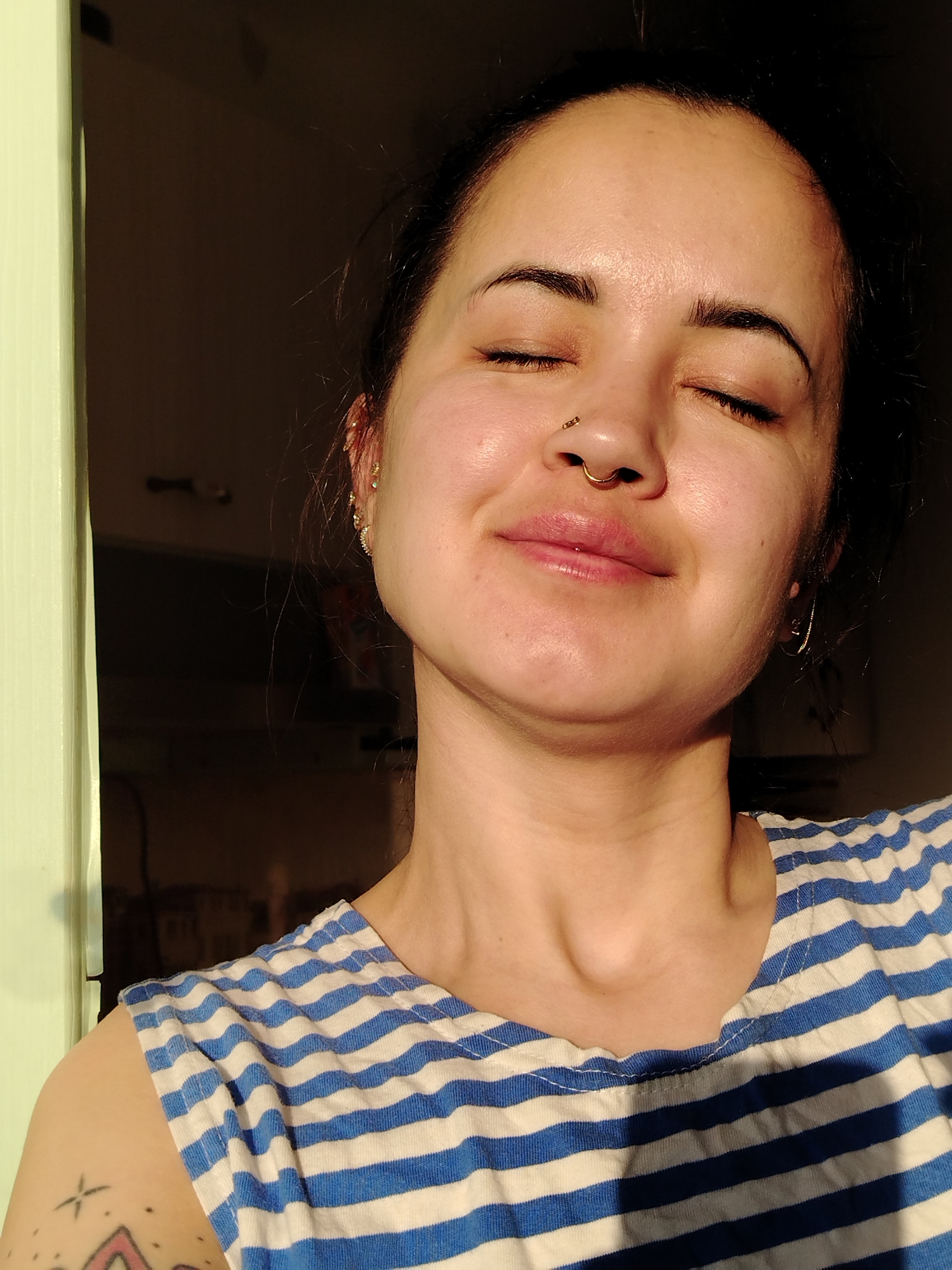 A medium close-up shot depicts a woman with her eyes closed and a serene smile, illuminated by strong, warm sunlight. She has dark hair pulled back and is wearing a blue and white striped shirt. She has a nose ring and earrings. Visible on her upper arm is a tattoo. The background is blurred, showing a doorframe and what appears to be an interior space. There is no evidence to definitively identify the location as Noviy, Russia.