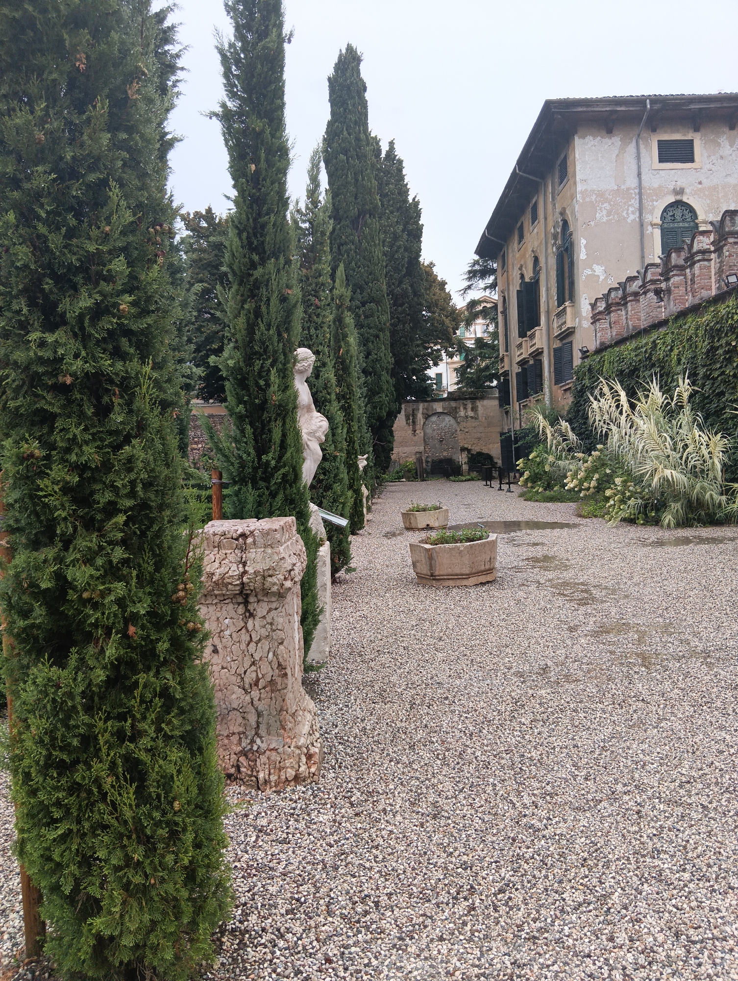 This is a full shot of a gravel courtyard in Verona, Italy. The courtyard is lined with tall evergreen trees on the left and a building on the right. The building appears to be an old villa with pale yellow stucco walls, dark green shutters, and a red tiled roof. A section of the building's wall is adorned with ivy. In the foreground, there are two large, weathered stone planters, one of which contains some sparse greenery. The gravel path shows evidence of recent rain, with puddles reflecting the overcast sky. In the middle ground, a classical statue of a woman is visible between the trees, and a large, rough-hewn stone pillar stands in the immediate foreground. The scene is tranquil and evokes a sense of historical grandeur.