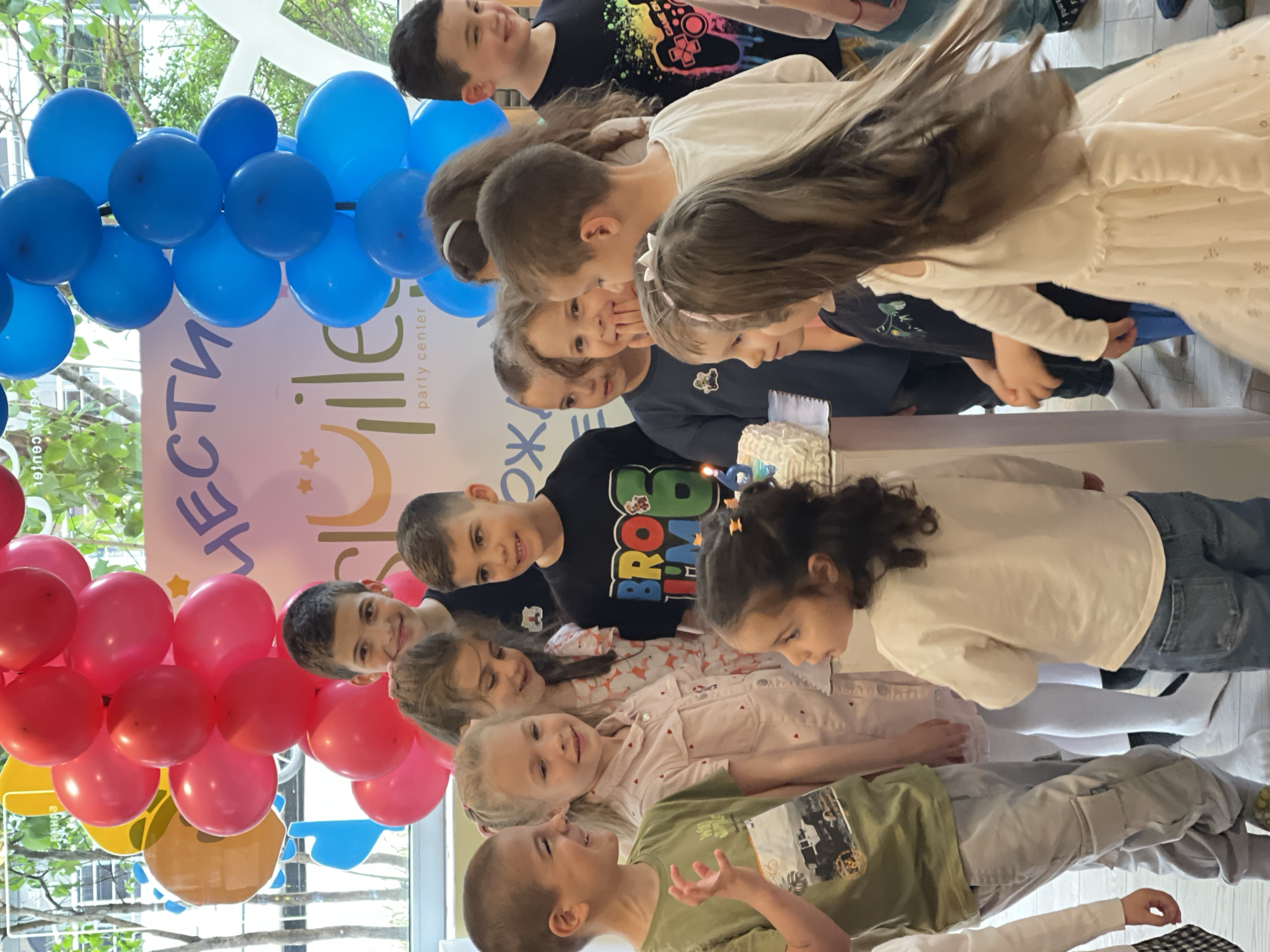 A group of children are gathered around a birthday cake in what appears to be a party center. The setting is brightly lit with natural light streaming in through a large window. There are decorations including an arch of red and blue balloons on the left side of the frame. A backdrop featuring text in Bulgarian is behind the children, indicating that the location is most likely in Bulgaria.
The children, about a dozen in total, are of varying ages and are dressed in casual clothing. They are positioned around a table with a cake. They appear to be anticipating or celebrating a birthday. Some of them are smiling and looking at the cake, while others are looking off to the side, either lost in thought or observing someone outside of the camera's view.
The event seems lively and cheerful, suggesting a festive occasion. The environment seems safe, family-friendly and festive. While the text on the backdrop indicates it is a “party center,” its location within Sofia specifically is unknown.