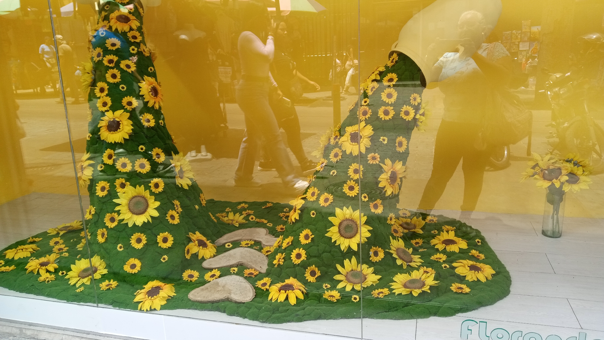 The image shows a storefront display in Unknown, Unknown. The display features an arrangement resembling a waterfall of sunflowers flowing out of two white containers or vases. The base is covered in green moss, with scattered sunflowers and a few wood-like stepping stones. A vase with sunflowers is located to the right. Reflections on the window show pedestrians and a motorcycle, blurring the view into the store. The bottom right corner has a partial word that appears to be related to the store's name.