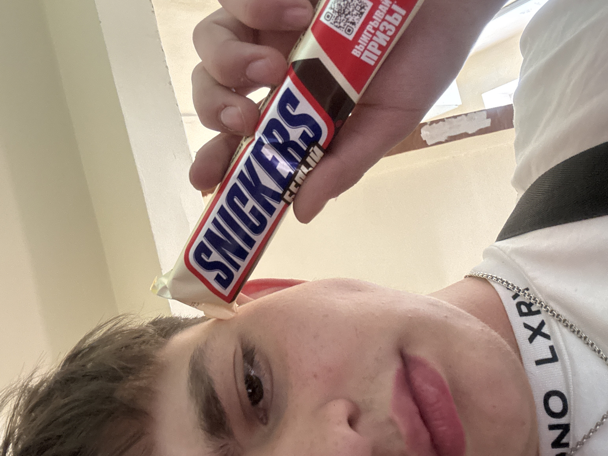 The image is a selfie taken from a low angle, focusing on a person's face with a Snickers bar balanced on their forehead. The person has fair skin, short brown hair, and is wearing a white shirt with black lettering and a silver chain necklace. The Snickers bar is in its packaging, featuring a red and white design with a QR code. The background is a plain, light-colored wall with a partially visible window frame. The location and city are unknown.