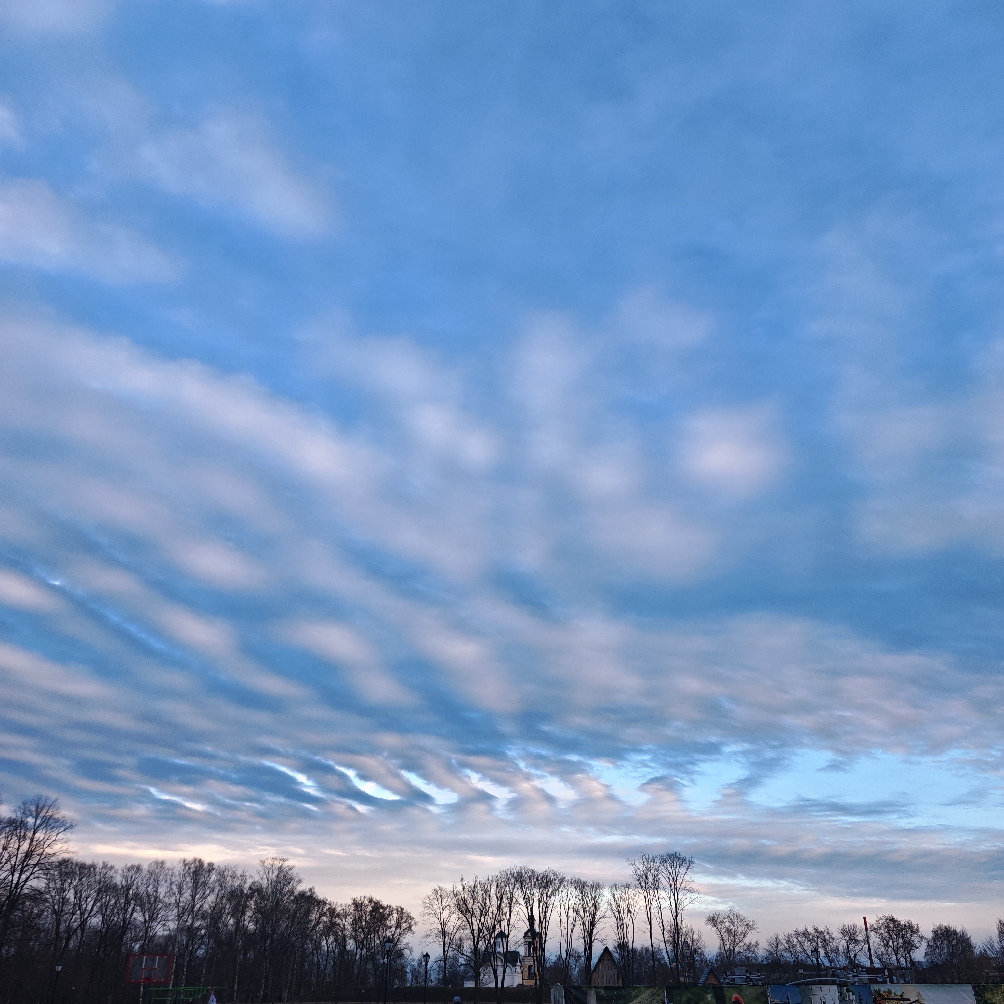 The image presents a view of the sky in Rostov, Russia. The sky is predominantly blue with streaks of elongated, slightly textured clouds. Below the sky, a line of bare trees stretches across the horizon, indicating either a winter or early spring season. In the middle of the trees, there is a church, and some buildings. In the bottom of the image, there are some objects, and a playground with no people visible. The environment seems to be a park or open space within the city of Rostov. There is no evidence of direct human activity or interaction in the frame. The lighting suggests that the picture was taken in the morning or evening.