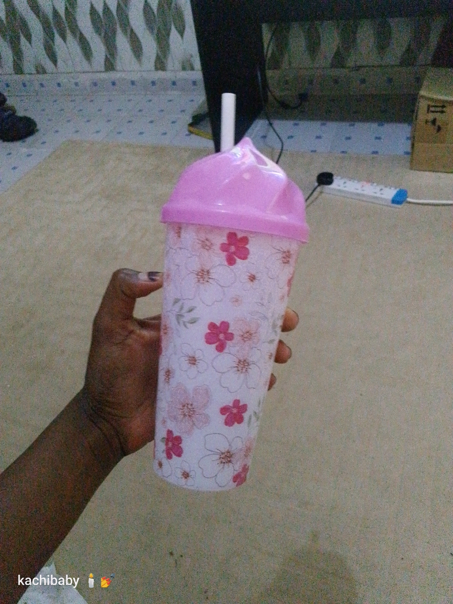 A person's hand is holding a tall, pink-lidded tumbler with a floral pattern. The tumbler is positioned in the foreground, occupying the majority of the right side of the image. The hand is dark-skinned and appears to be gently cupping the tumbler. The tumbler itself has a white base with a pattern of pink and white flowers. A white straw is visible emerging from the pink lid.
In the background, the environment appears to be an indoor space with a textured floor. There is a power strip with blue and white accents visible on a surface behind the tumbler, and a brown cardboard box to the far right. The wall in the far background has a patterned design. The lighting suggests an indoor setting. The watermark "kachibaby" with a candle and fireworks emoji is present in the bottom left corner. The image appears to be taken in Mtopanga ward, Kenya.