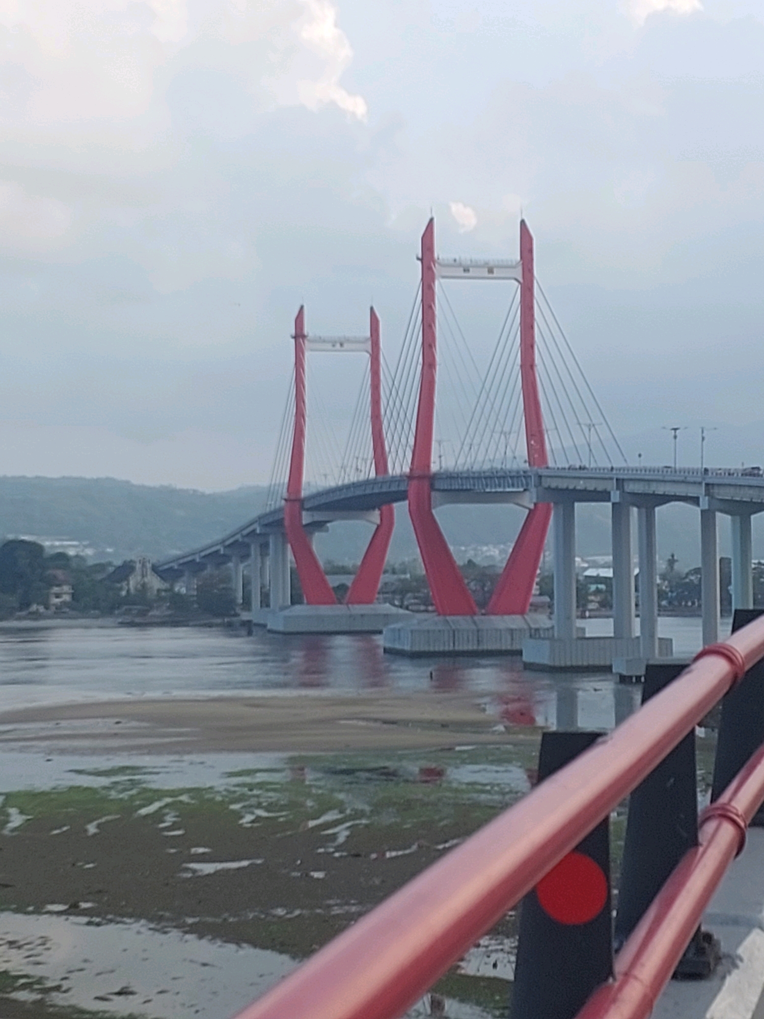The image depicts the Youtefa Bridge, a cable-stayed bridge in Ambon, Indonesia. The bridge features prominent red pylons supporting the roadway. The water below the bridge appears shallow, with exposed sand and patches of green vegetation, suggesting a tidal area or a low tide. In the background, a hilly landscape with scattered buildings and trees can be seen. The sky is overcast with some wispy clouds. In the foreground, a red railing with black vertical supports is visible, indicating the vantage point is likely from another bridge or elevated structure. There are no discernible people or ongoing actions in the scene, presenting a static view of the bridge and its surroundings.