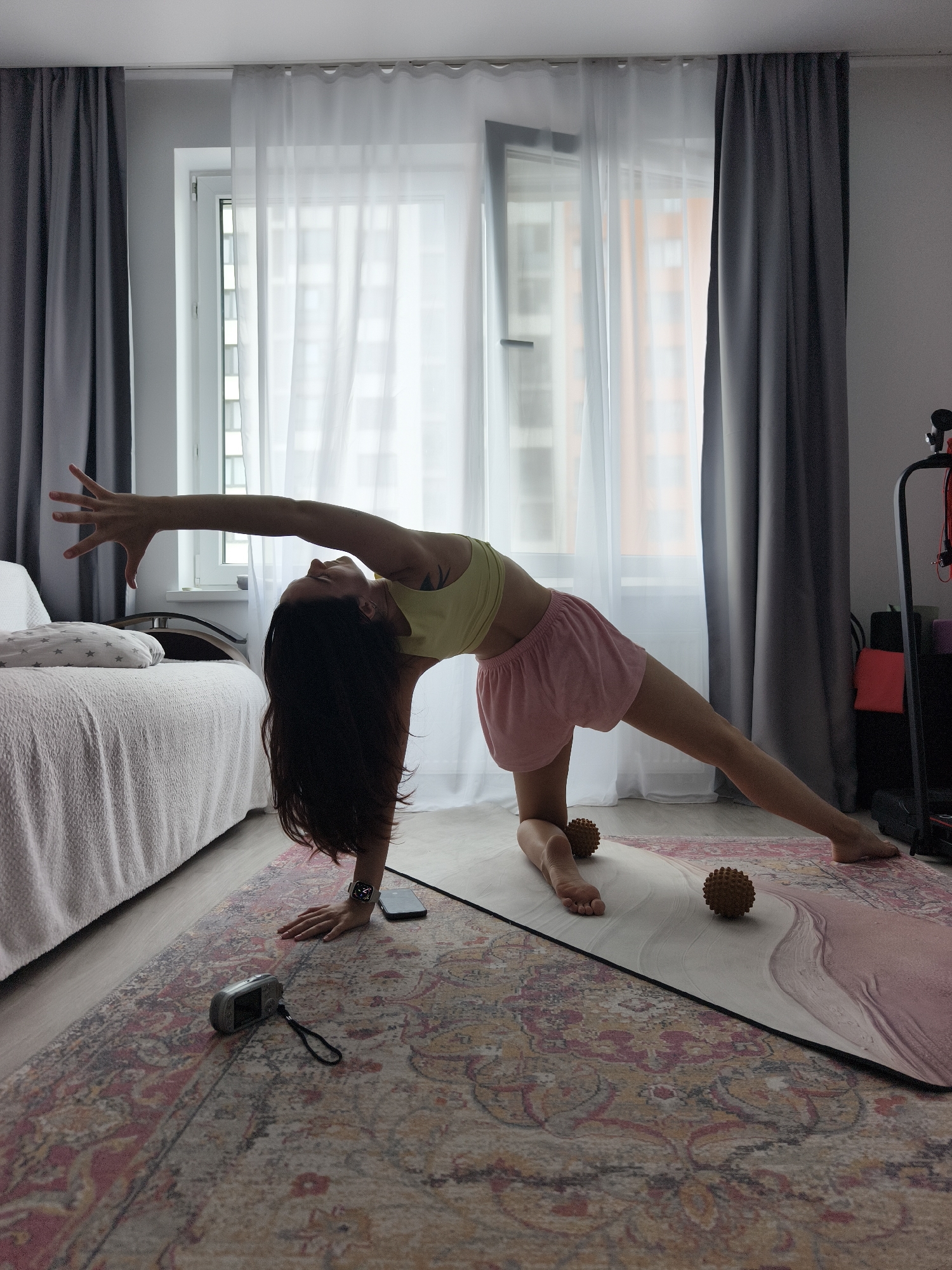 An adult female with long dark hair is depicted performing a yoga stretch indoors in a residential apartment in Tyumen, Russia. She is on a pink yoga mat, positioned over a large patterned Persian-style rug, kneeling on her left knee with her left hand on the mat, and her right leg extended to the side. Her left arm is extended upwards and arched back, with her head tilted to look up. She wears a light yellow crop top and pink athletic shorts. A smartwatch is visible on her left wrist. Two spiky brown massage balls are on the yoga mat. A black smartphone and a silver compact digital camera with a black strap are on the rug near her left arm. To the left, a bed with a white textured duvet and a gray star-patterned pillow is visible. In the background, a large window with white sheer curtains and dark gray opaque curtains reveals a blurry view of other apartment buildings. To the far right, black home gym equipment with red resistance bands and various yoga blocks are partially visible.